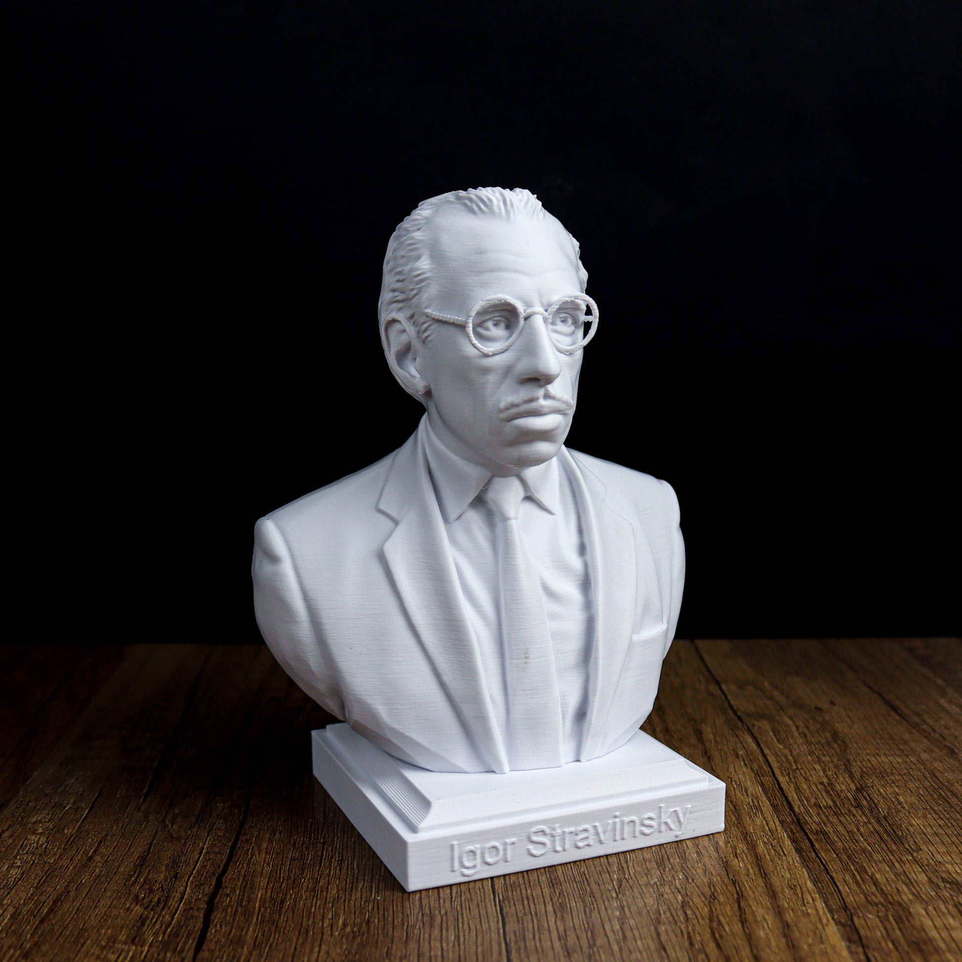 A white bust of a man wearing glasses and a suit, sitting on a wooden surface.