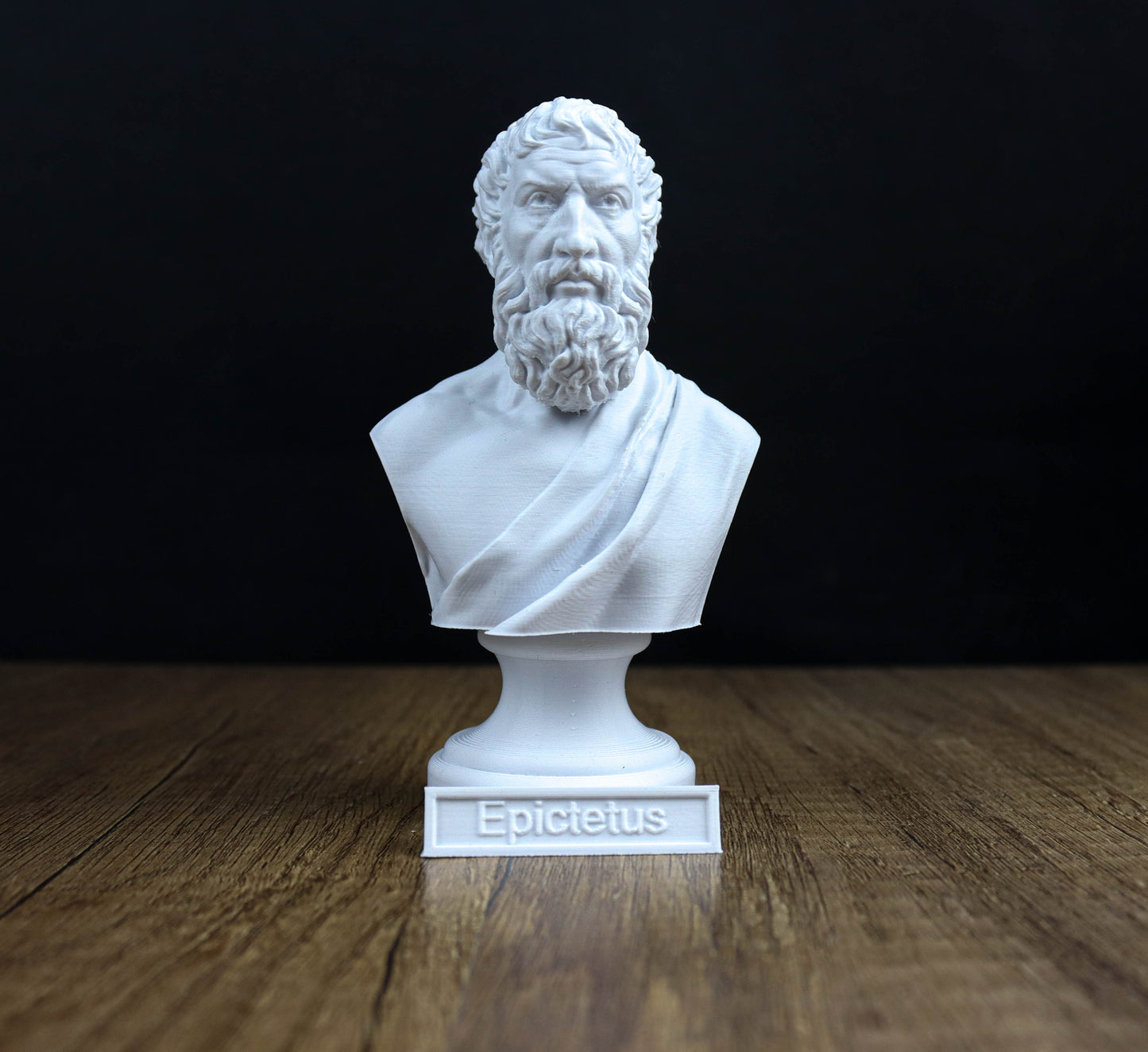 A white bust of a bearded man with a serious expression, likely depicting the ancient Greek philosopher Epicurus, is displayed on a wooden surface against a black background.