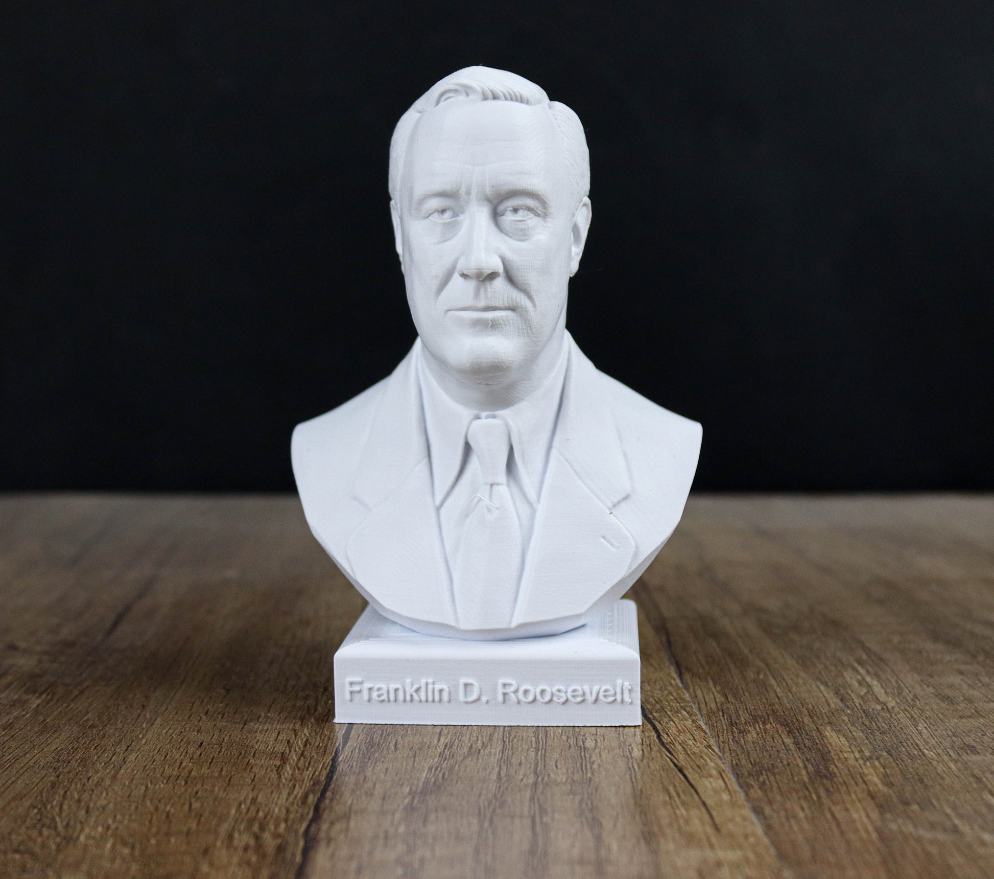 A white bust of a man, likely Franklin D. Roosevelt, is displayed on a wooden surface against a black background.