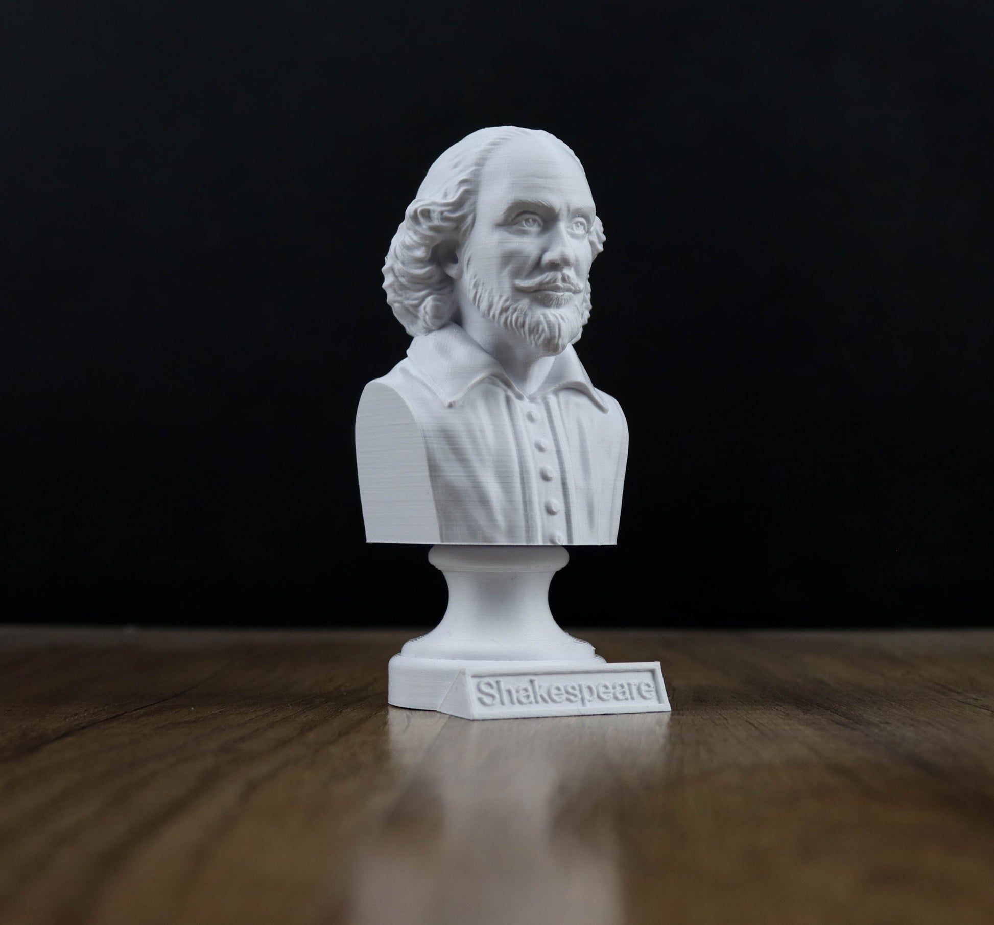 A white bust of William Shakespeare, a famous English playwright, is displayed on a wooden surface against a black background.