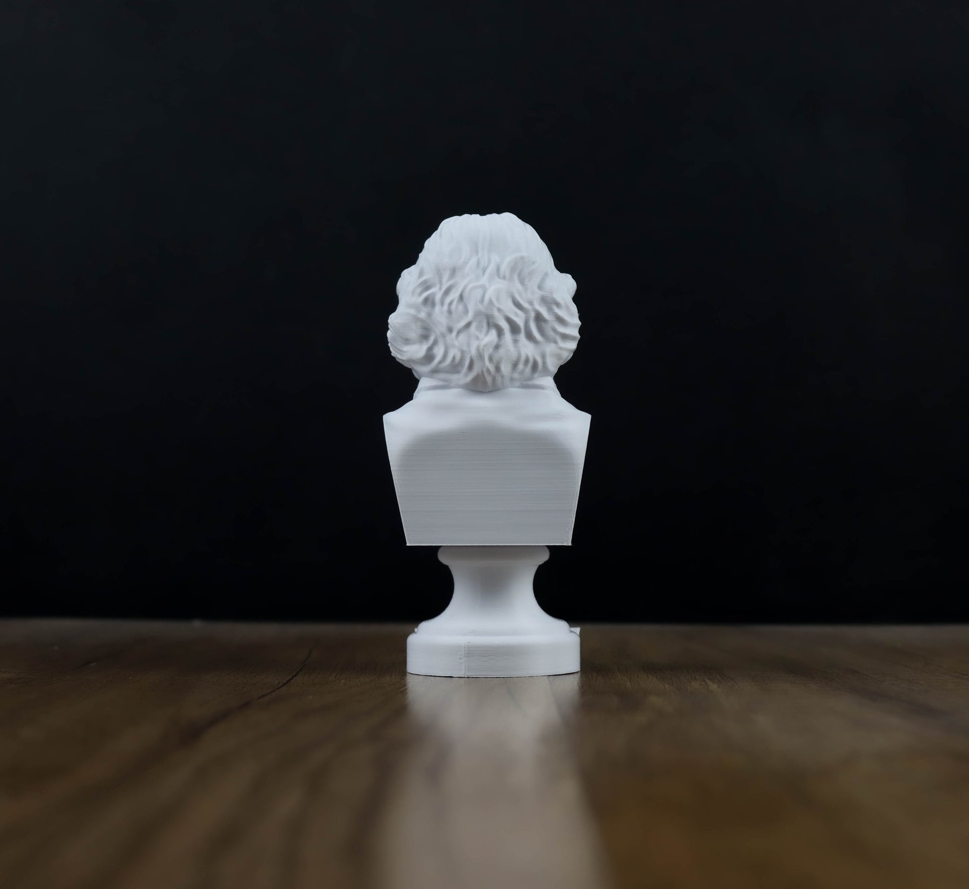 A white bust sculpture is displayed on a wooden surface against a dark background.
