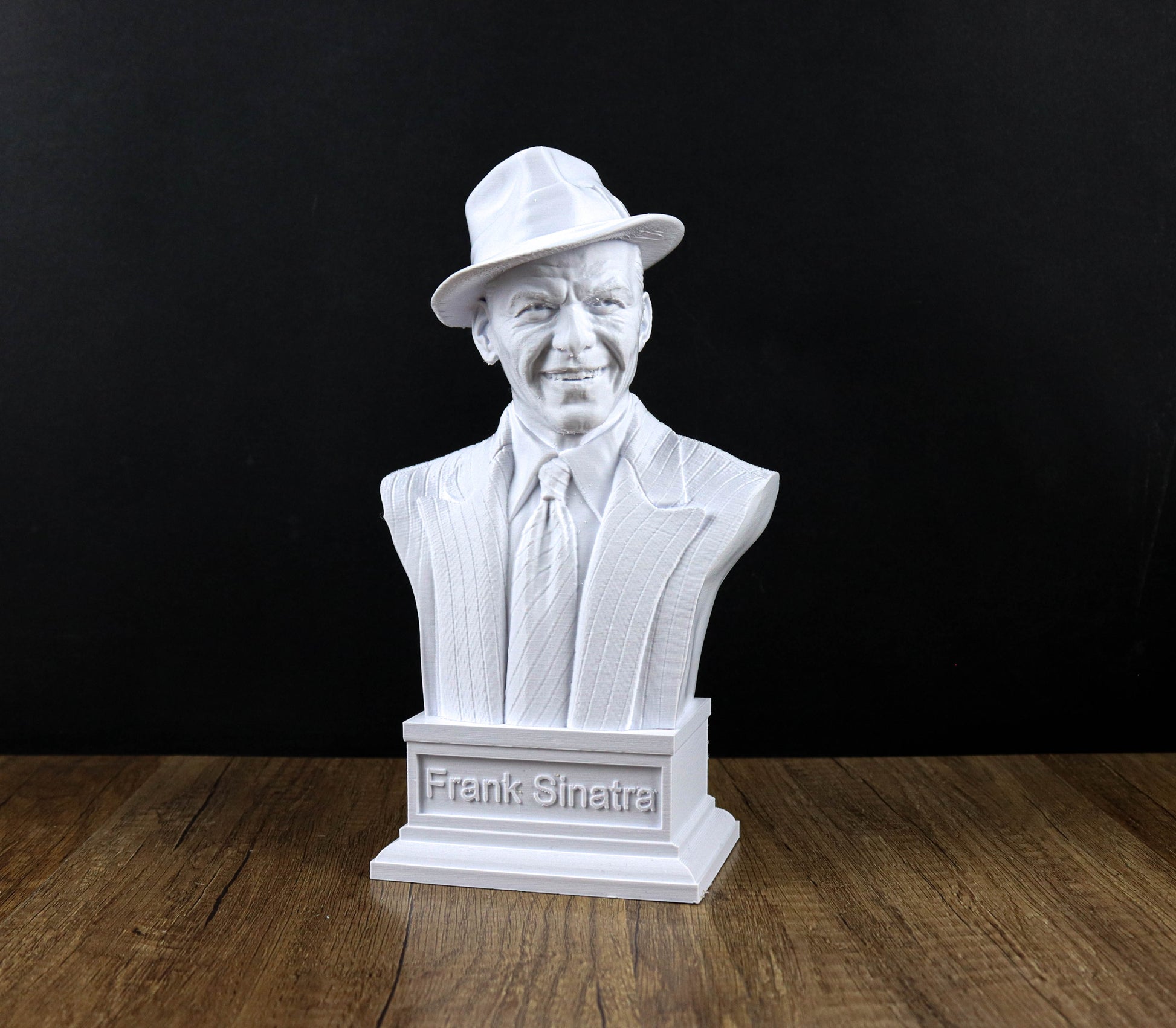 A white bust of Frank Sinatra, a famous American singer and composer, is displayed on a wooden surface against a black background.