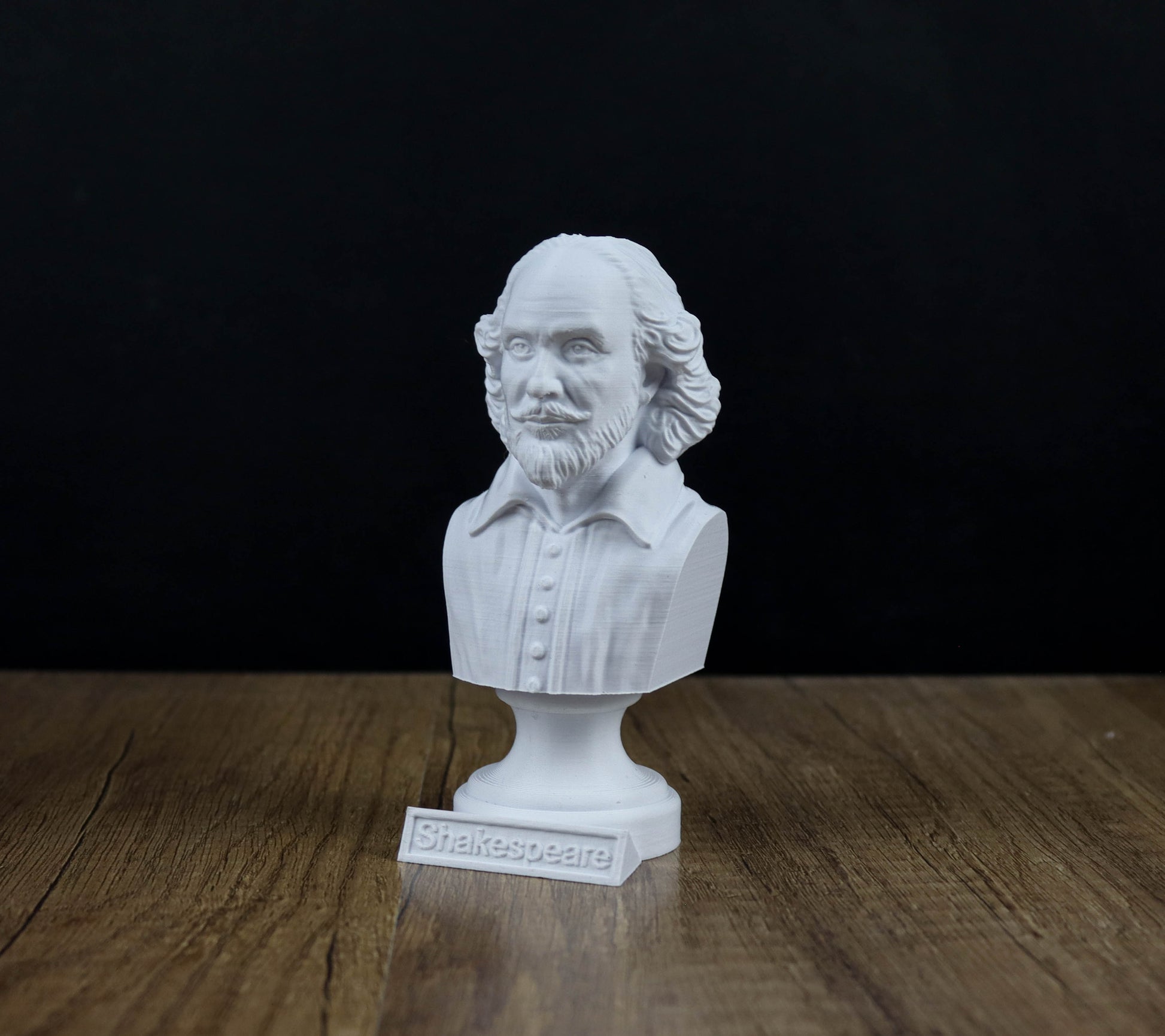 A white bust of William Shakespeare, a famous English playwright, is displayed on a wooden surface against a black background.