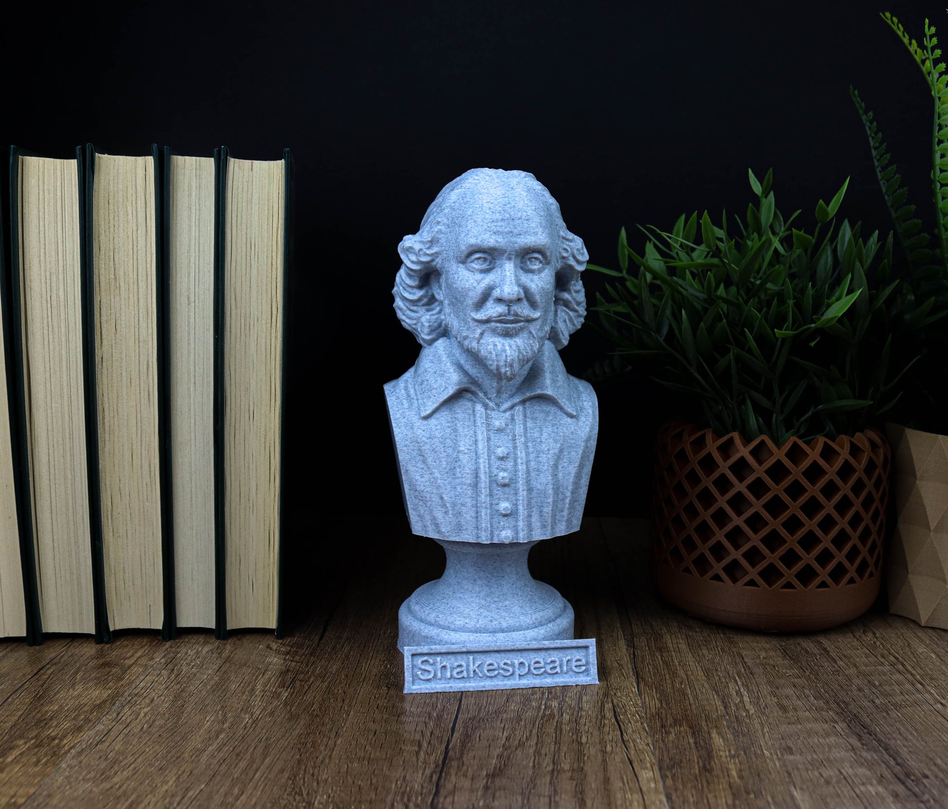 A bust of William Shakespeare is displayed on a wooden surface, surrounded by books and a potted plant.