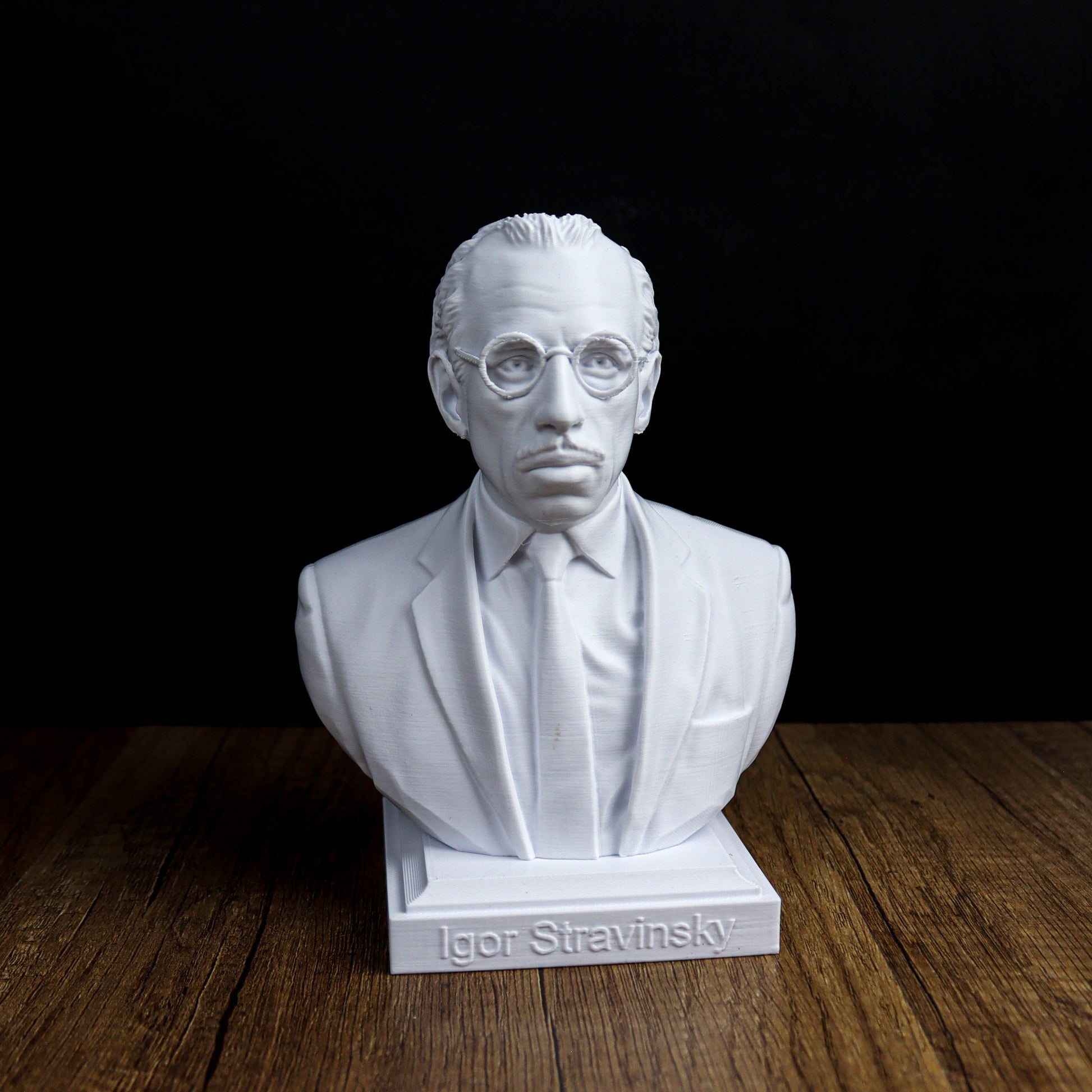 A white bust of a man wearing glasses and a suit, sitting on a wooden surface.