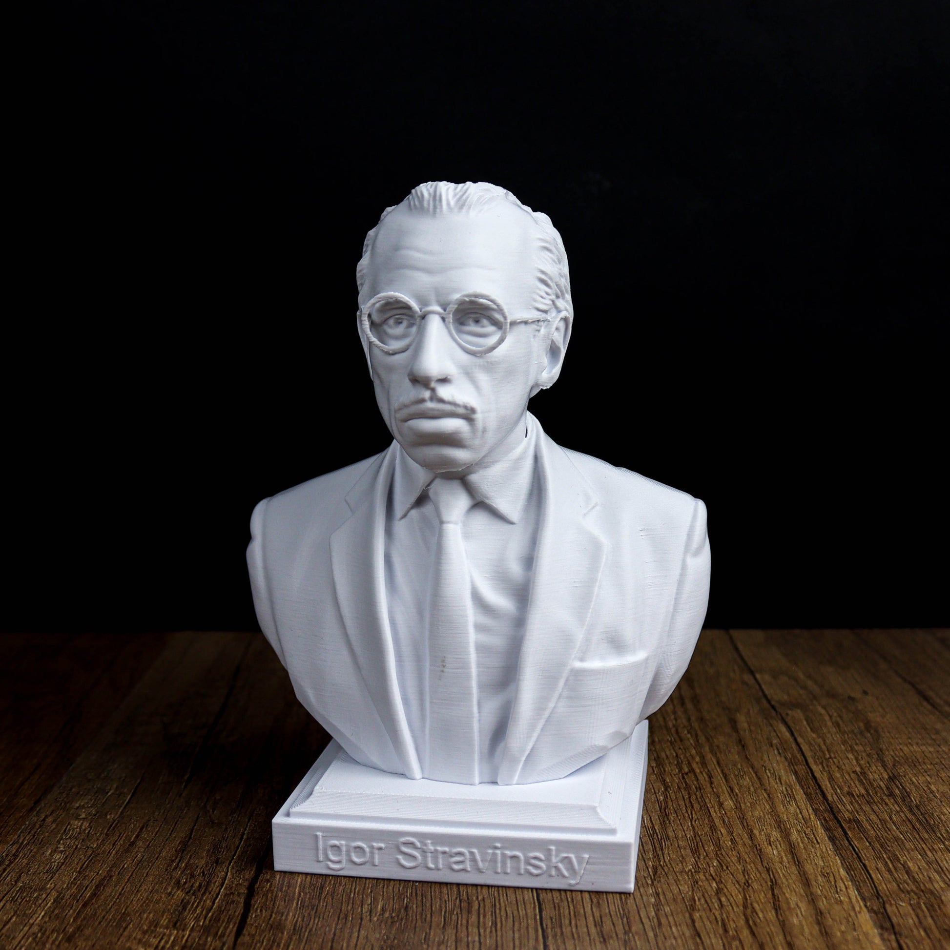 A white bust of a man wearing glasses and a suit, sitting on a wooden surface.