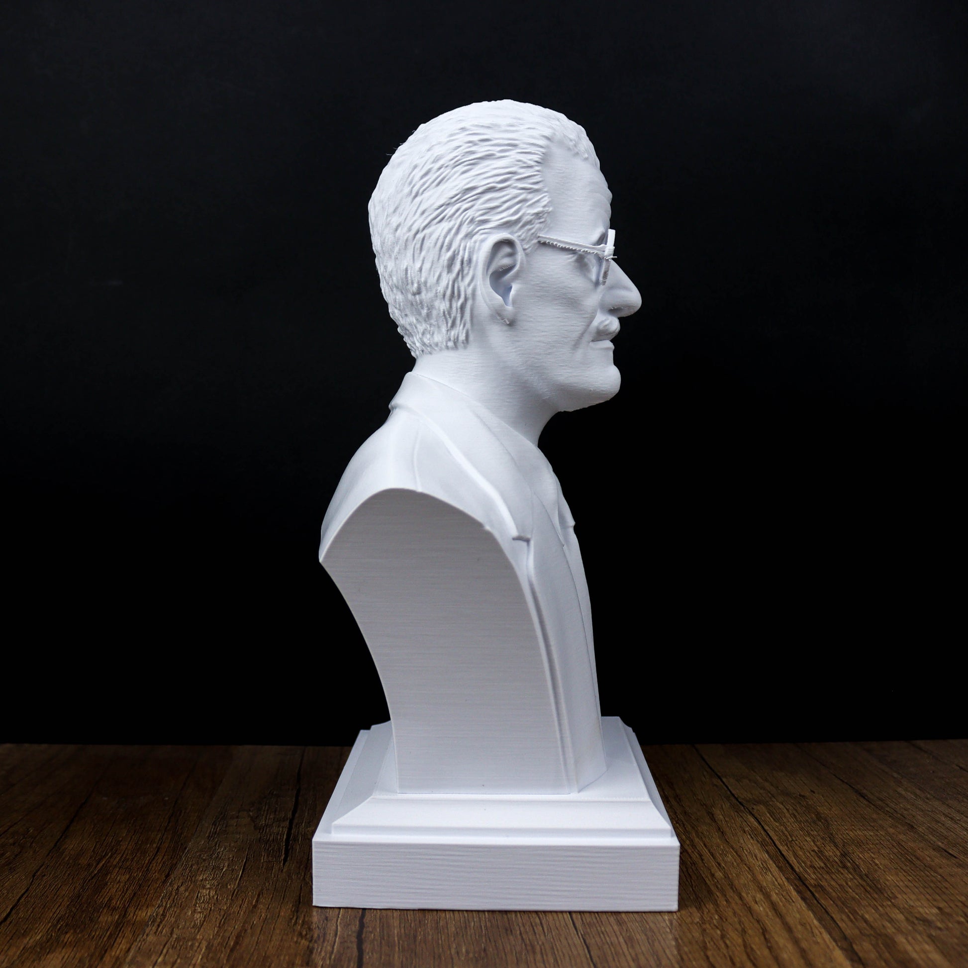 A white bust sculpture of a man wearing glasses is displayed on a wooden surface against a black background.