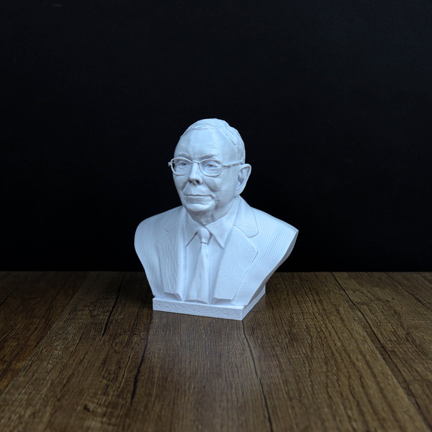A white bust of a man wearing glasses and a suit is displayed on a wooden surface against a black background.