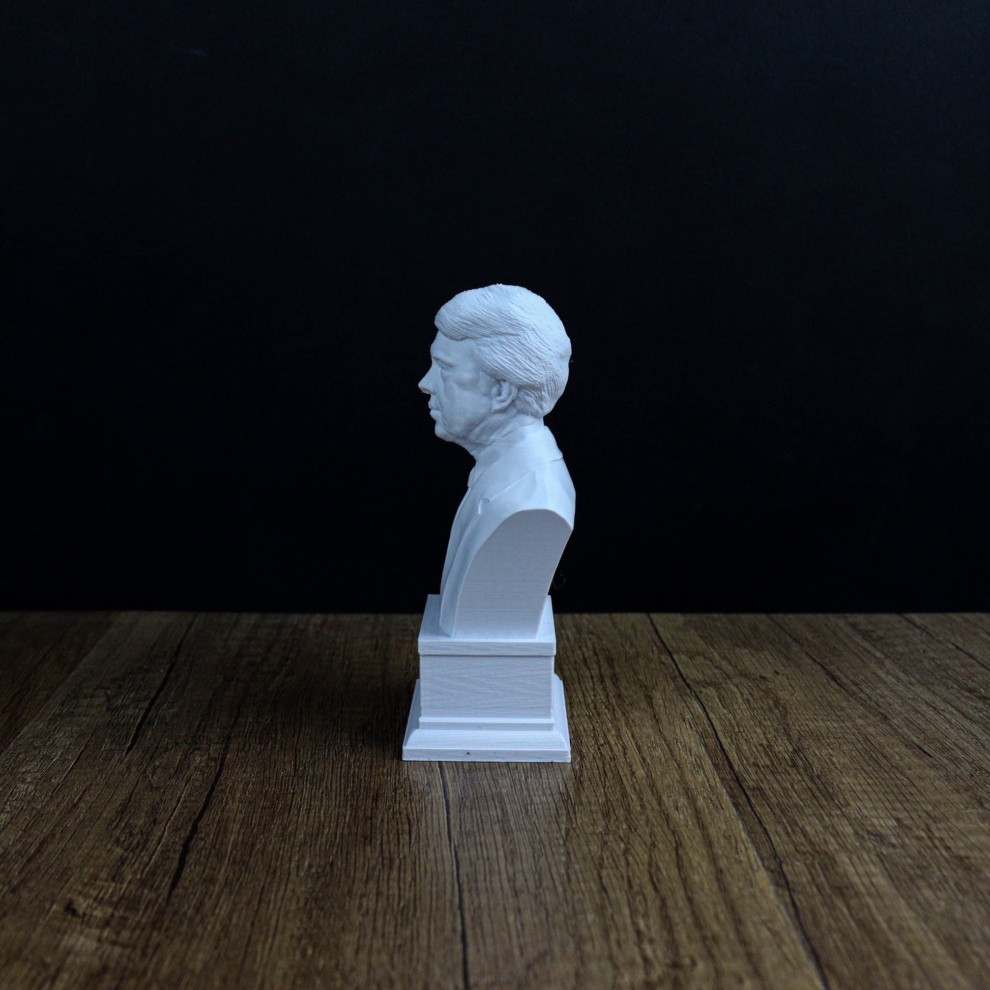 A white bust sculpture is displayed on a wooden surface against a dark background.