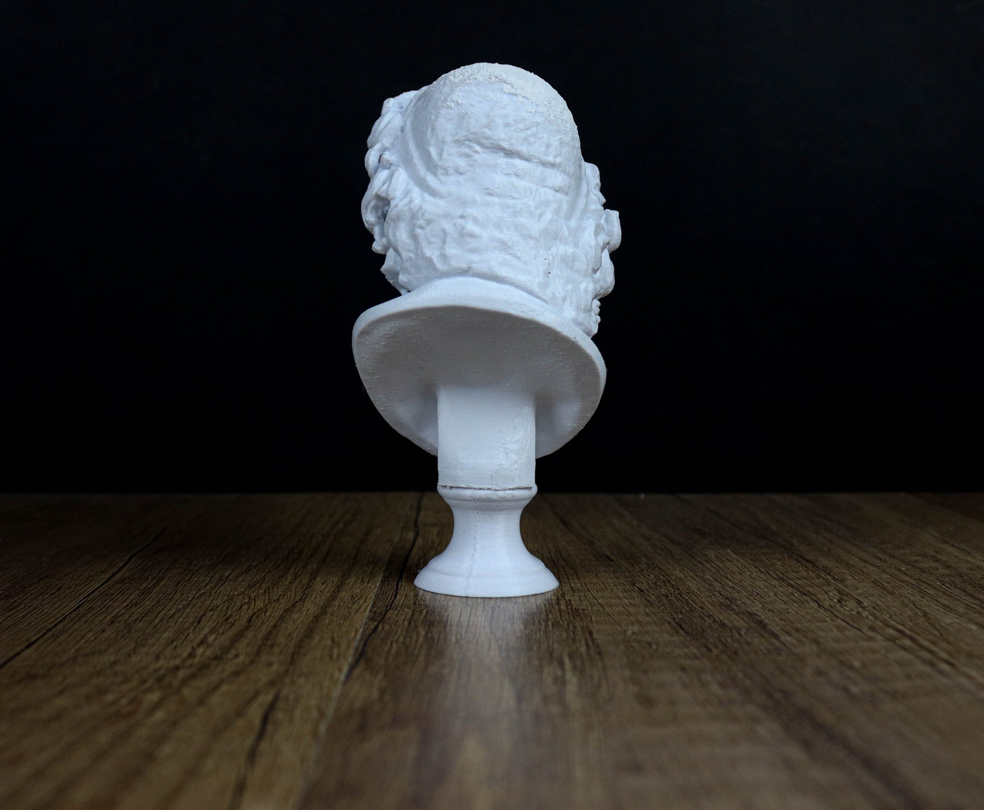 A white bust sculpture is displayed on a wooden surface against a dark background.