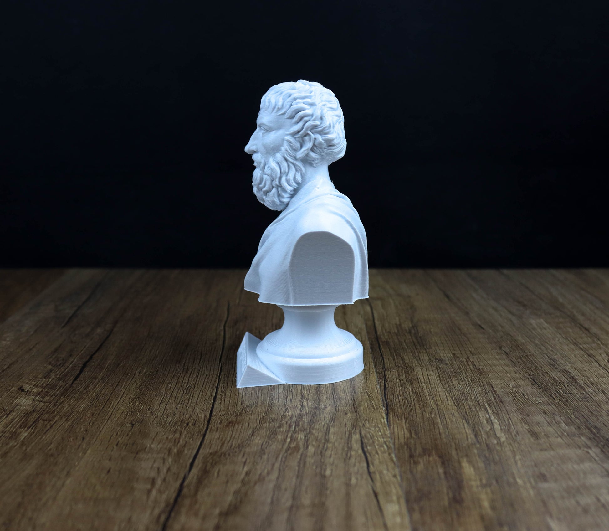 A white bust of a bearded man is displayed on a wooden surface against a dark background.