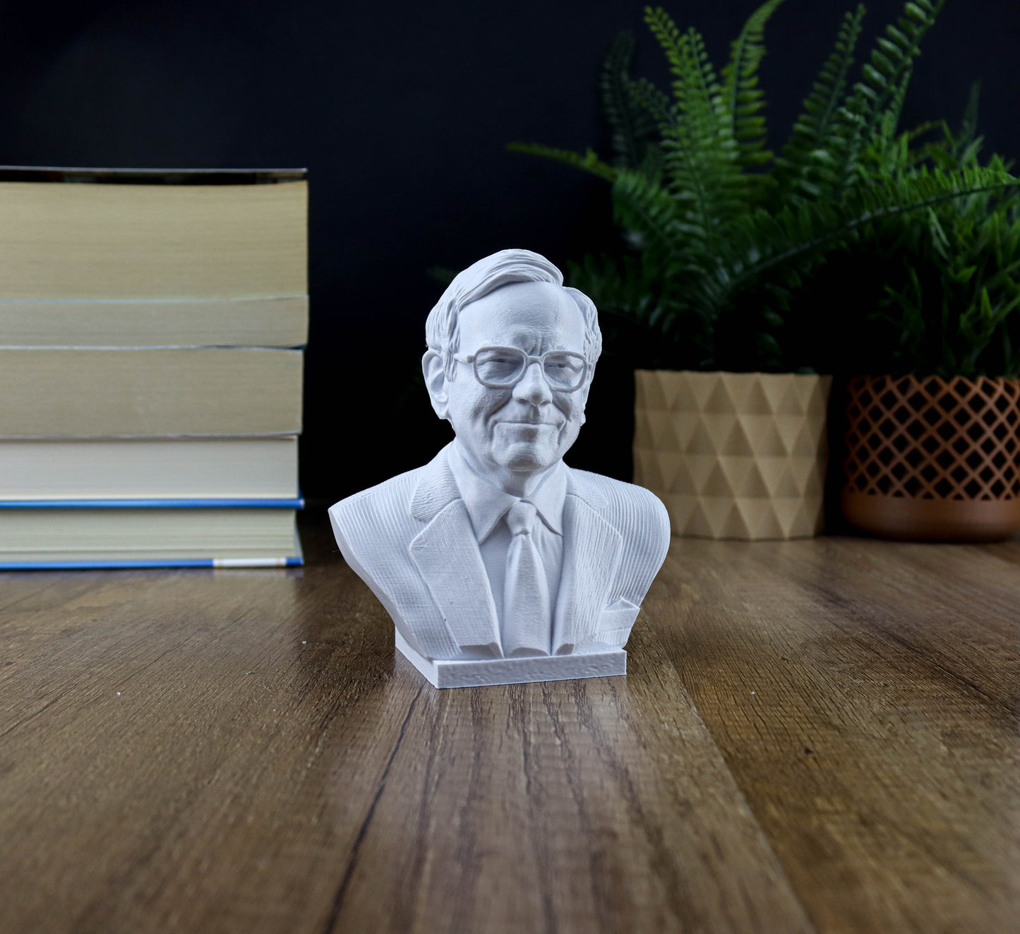 A white bust of a man wearing a suit and tie is displayed on a wooden surface, with a stack of books and a potted plant in the background.