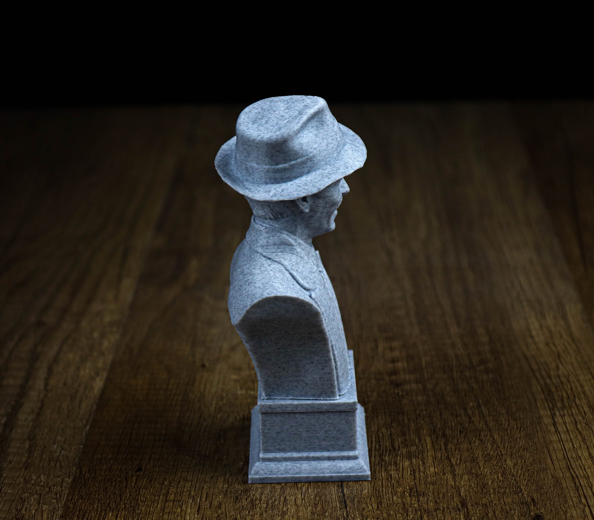 A small gray bust of a person wearing a hat, sitting on a wooden surface.