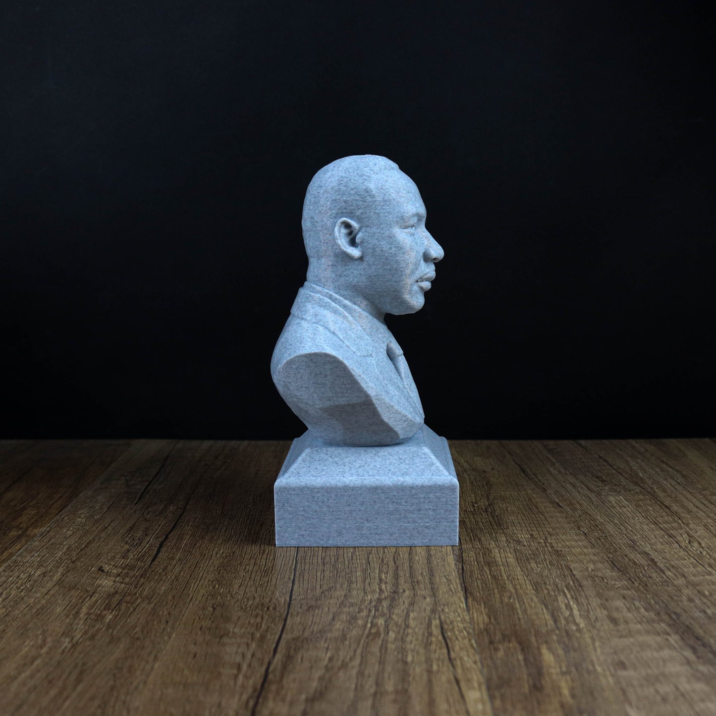 A gray bust of a person is sitting on a wooden surface against a black background.