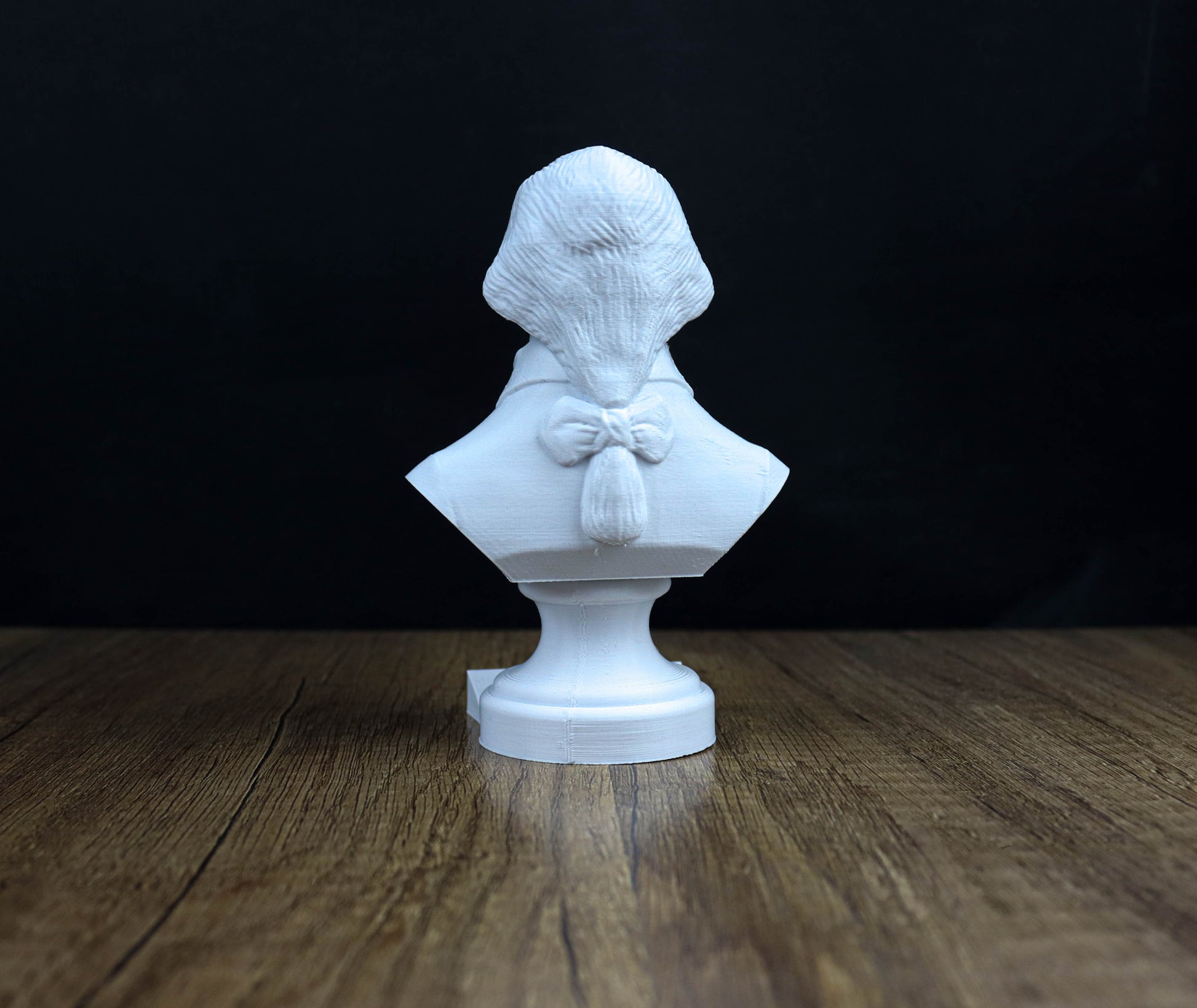 A white bust sculpture of a person is displayed on a wooden surface against a dark background.