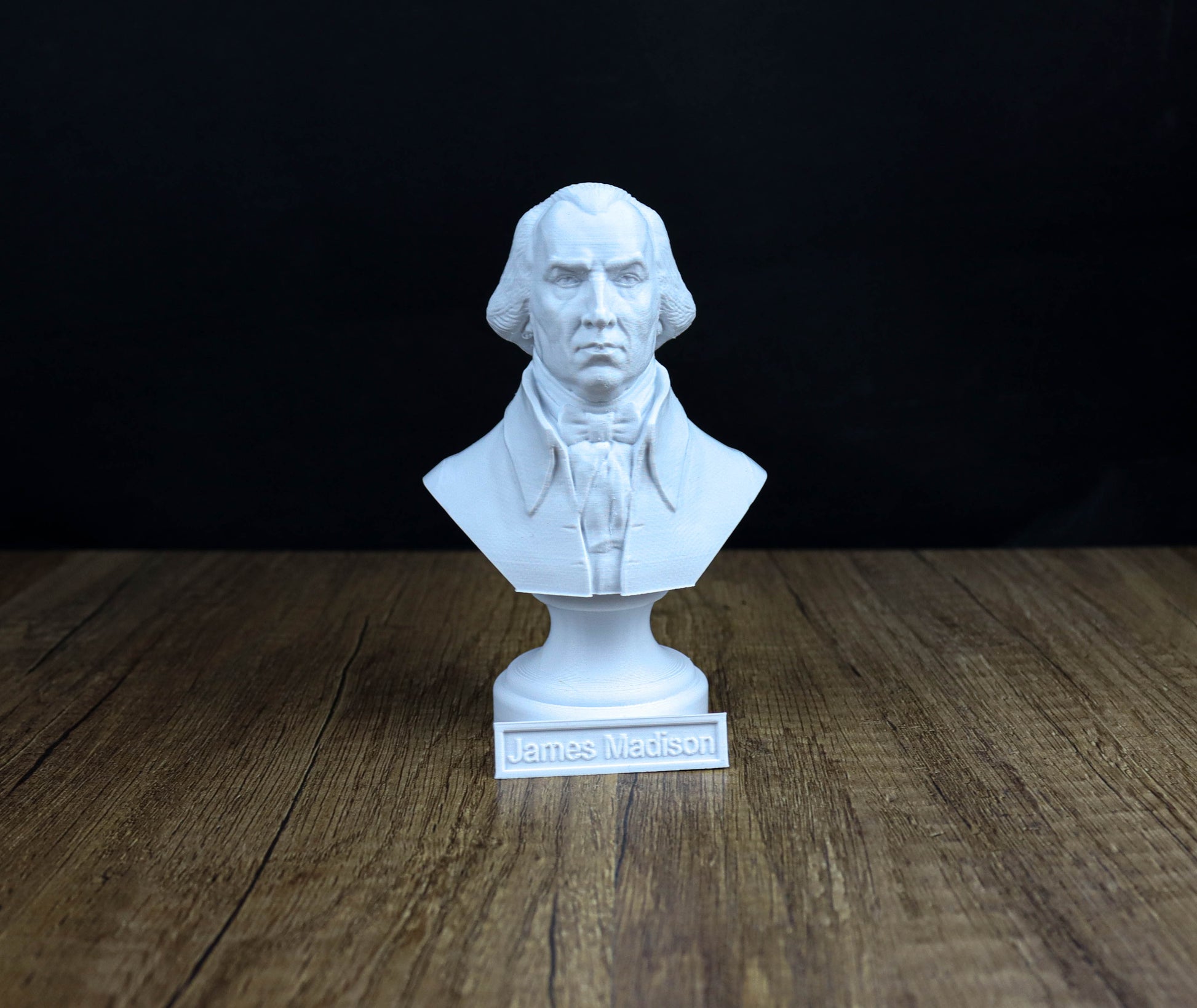 A white bust of a man, likely James Madison, is displayed on a wooden surface against a black background.