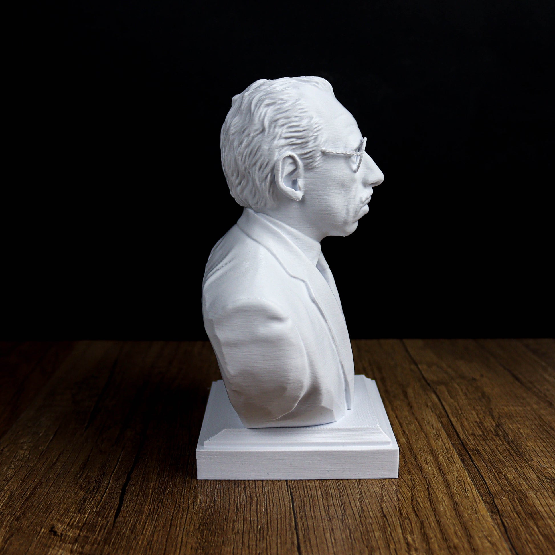 A white bust of a man wearing glasses is displayed on a wooden surface against a black background.