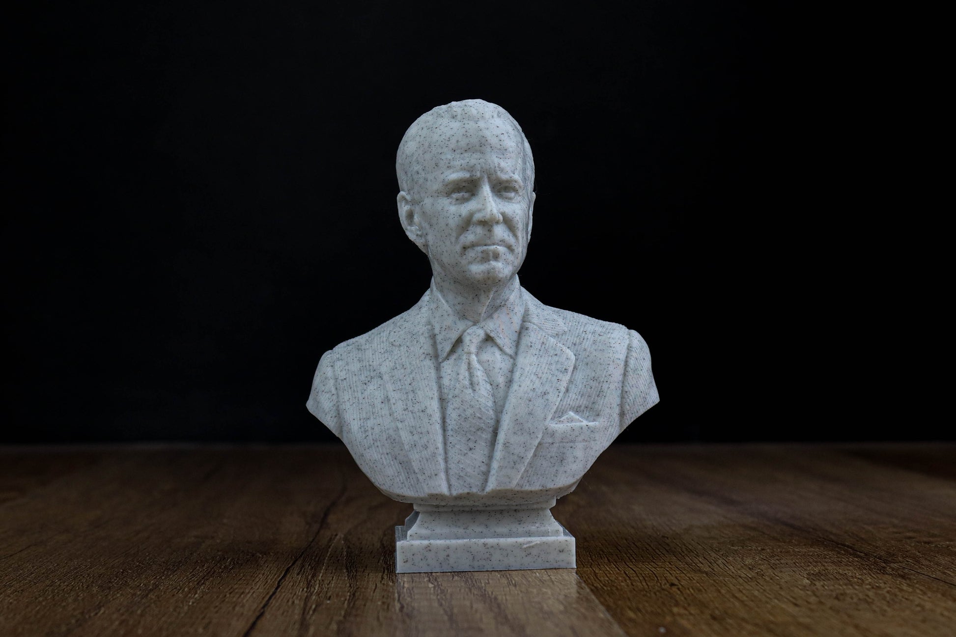 A white bust of a man wearing a suit and tie, sitting on a wooden surface against a black background.