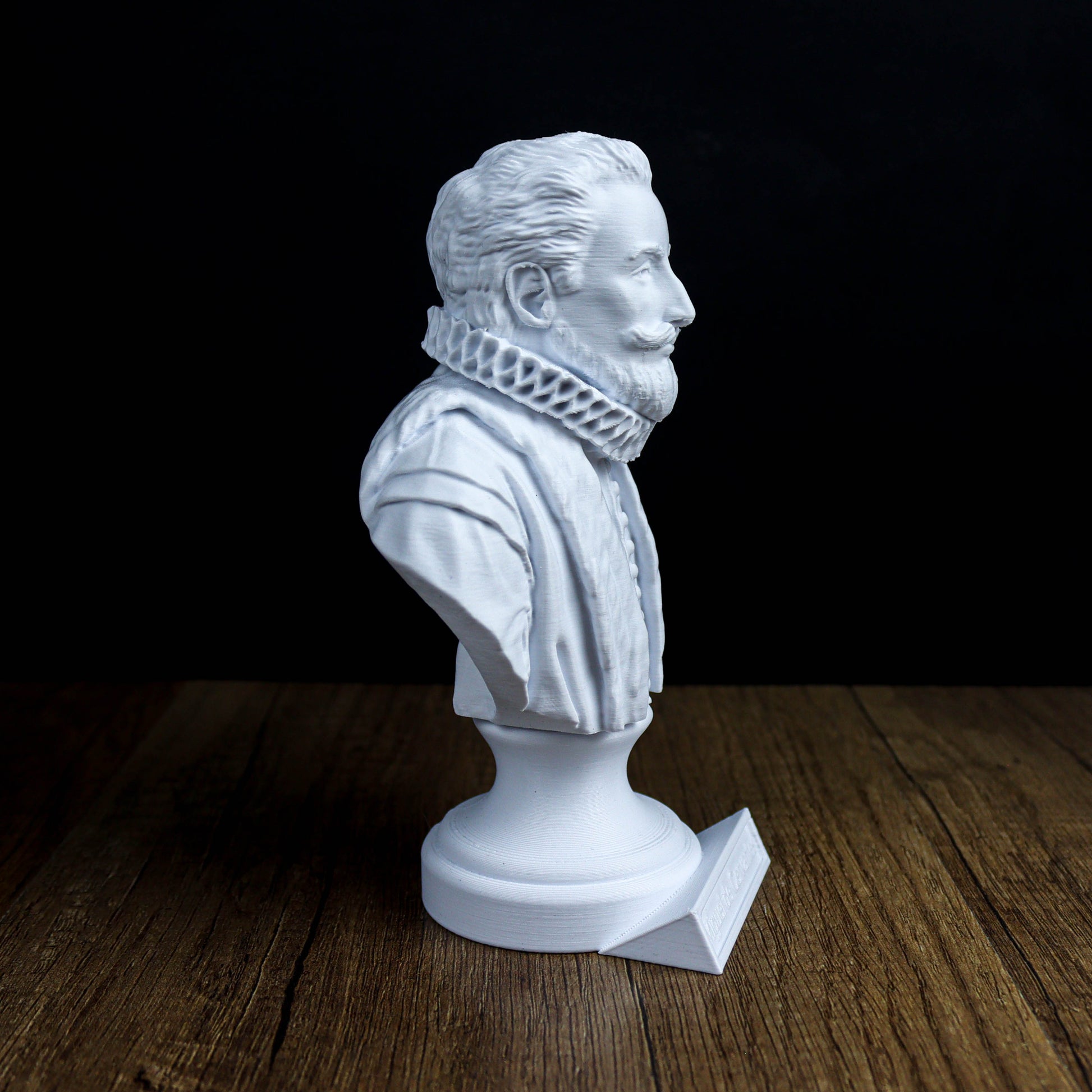 A white bust of a man with a beard and mustache, wearing a ruffled collar, is displayed on a wooden surface against a dark background.