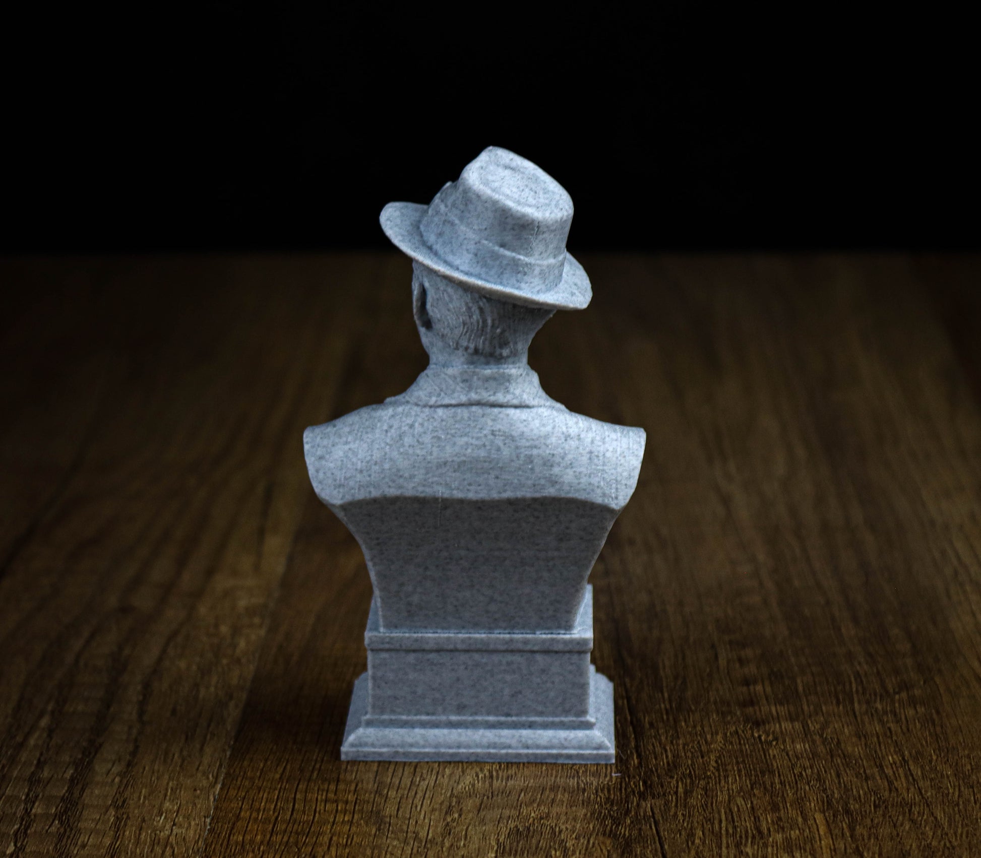 A gray stone statue of a man wearing a hat, sitting on a wooden surface.