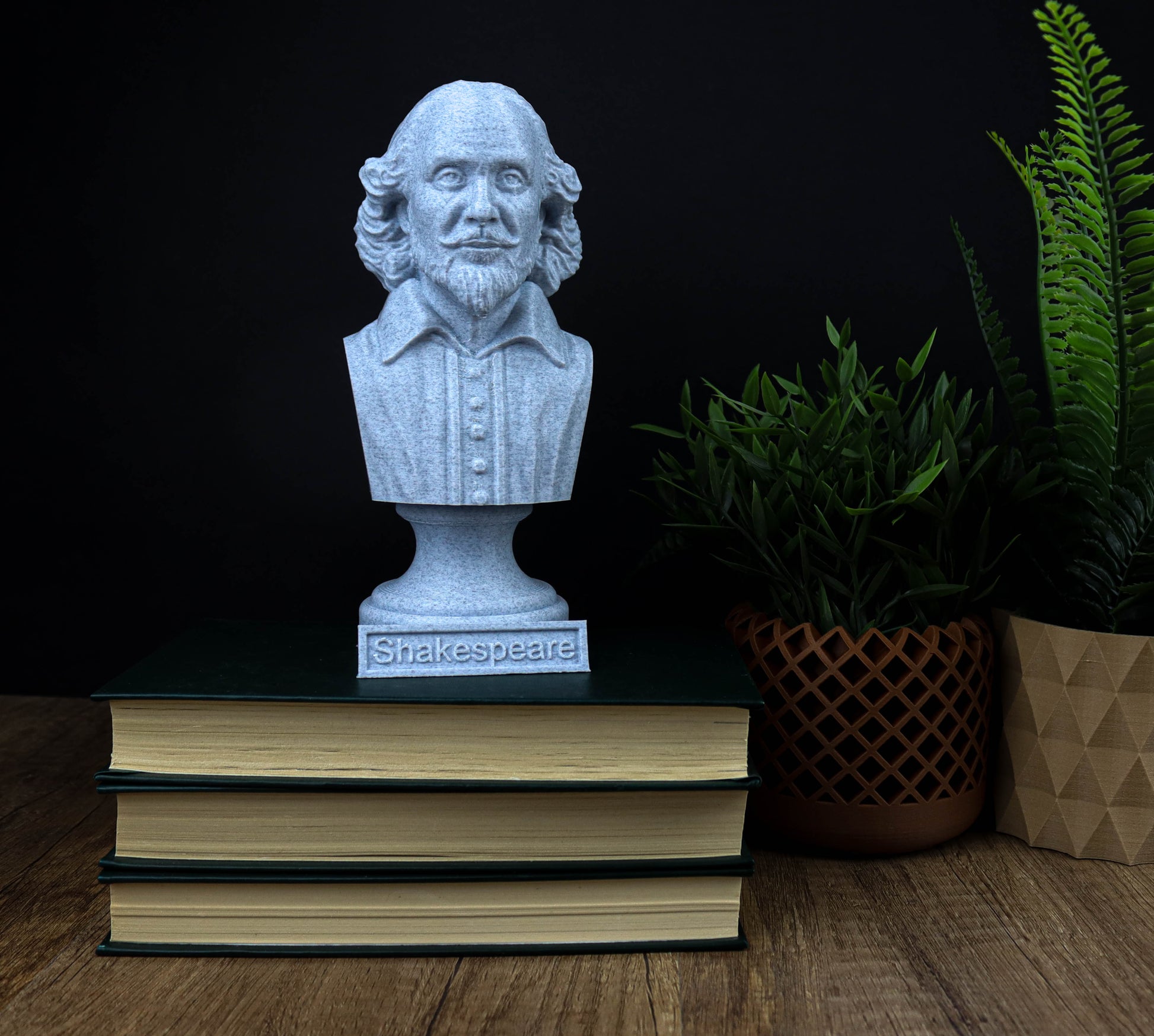A bust of William Shakespeare is displayed on top of a stack of books, with a potted plant and a geometric-patterned bag to the side.
