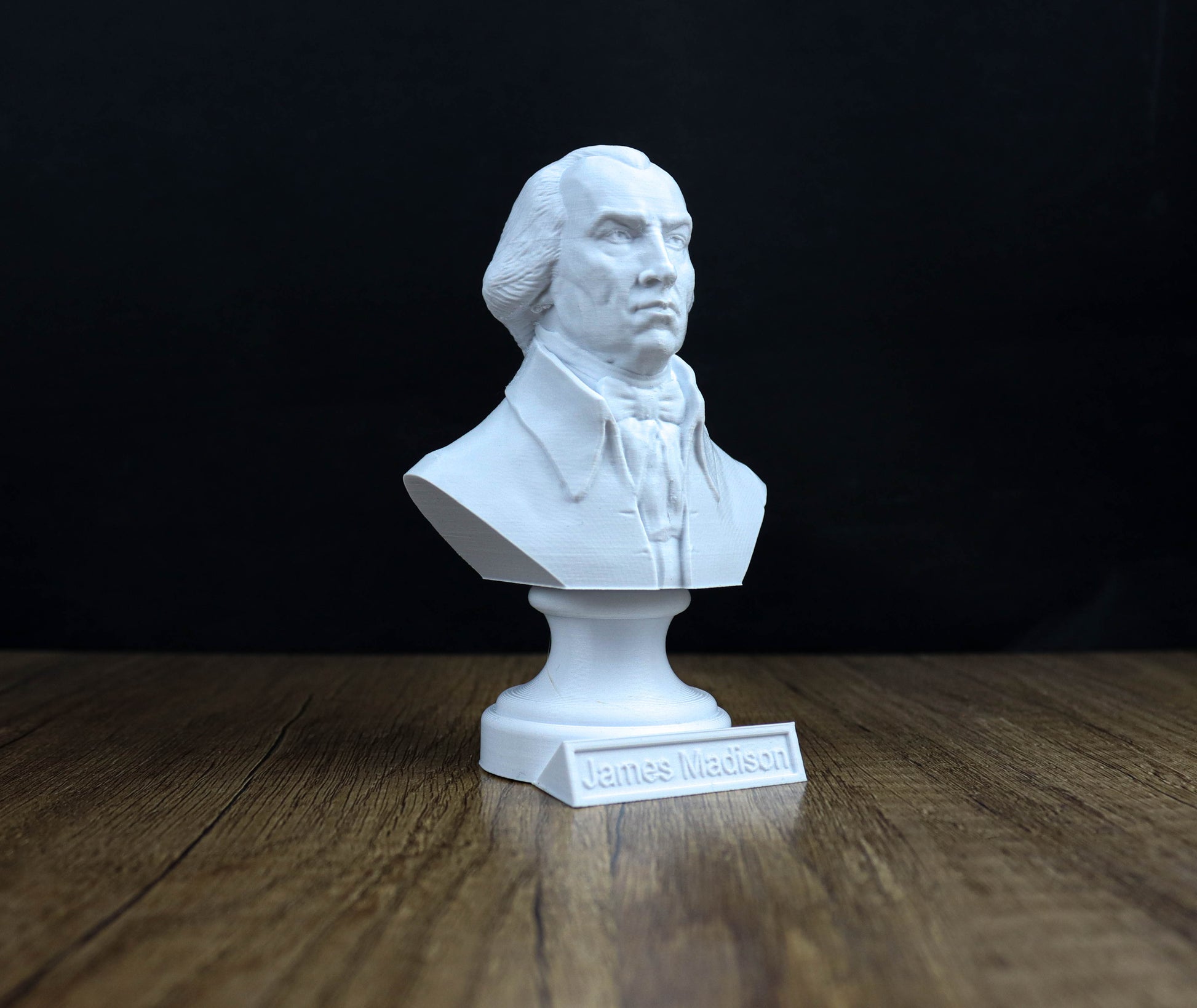 A white bust of a man, likely James Madison, is displayed on a wooden surface against a black background.