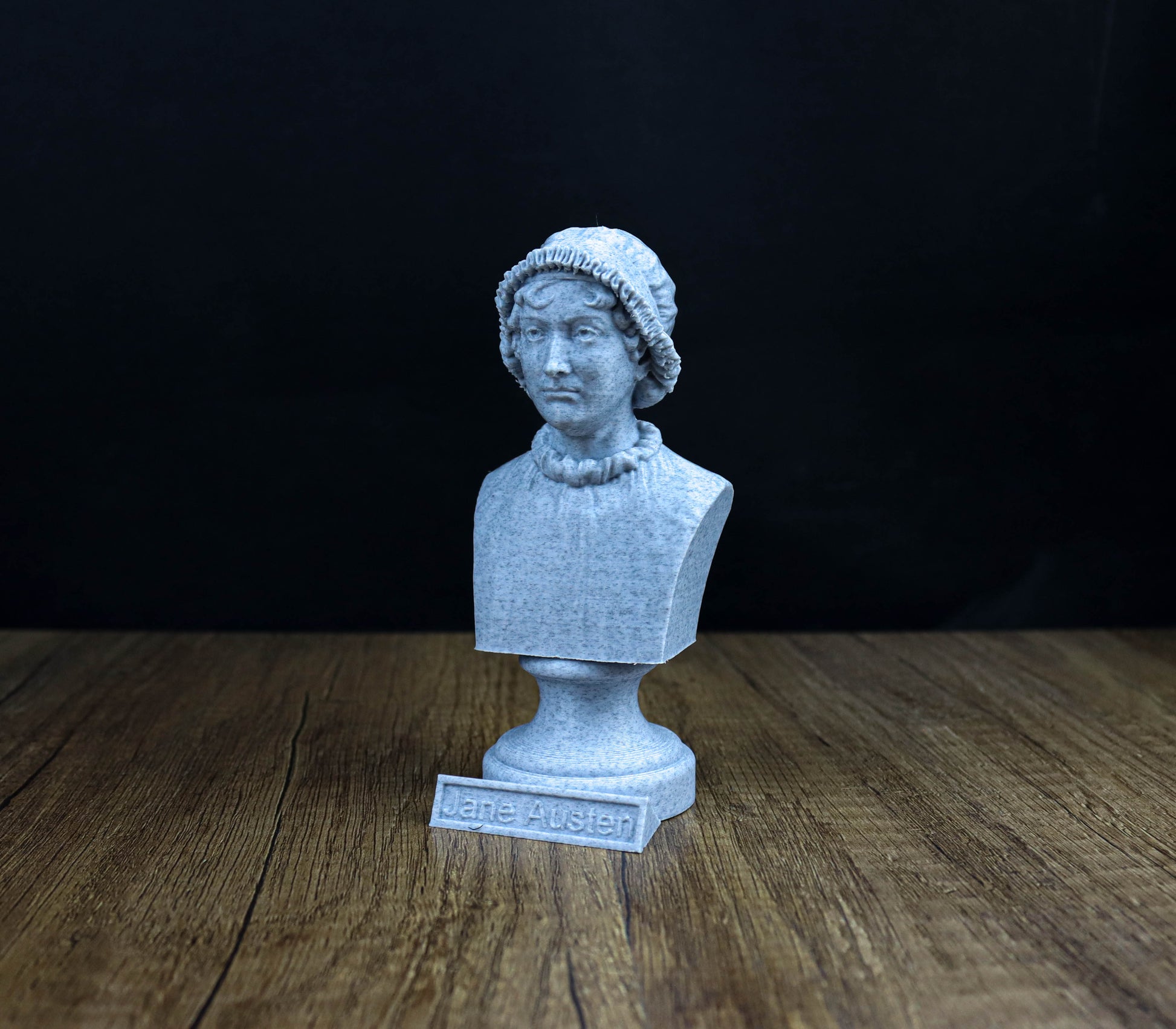 A small bust of a person, possibly a historical figure, is displayed on a wooden surface against a dark background.