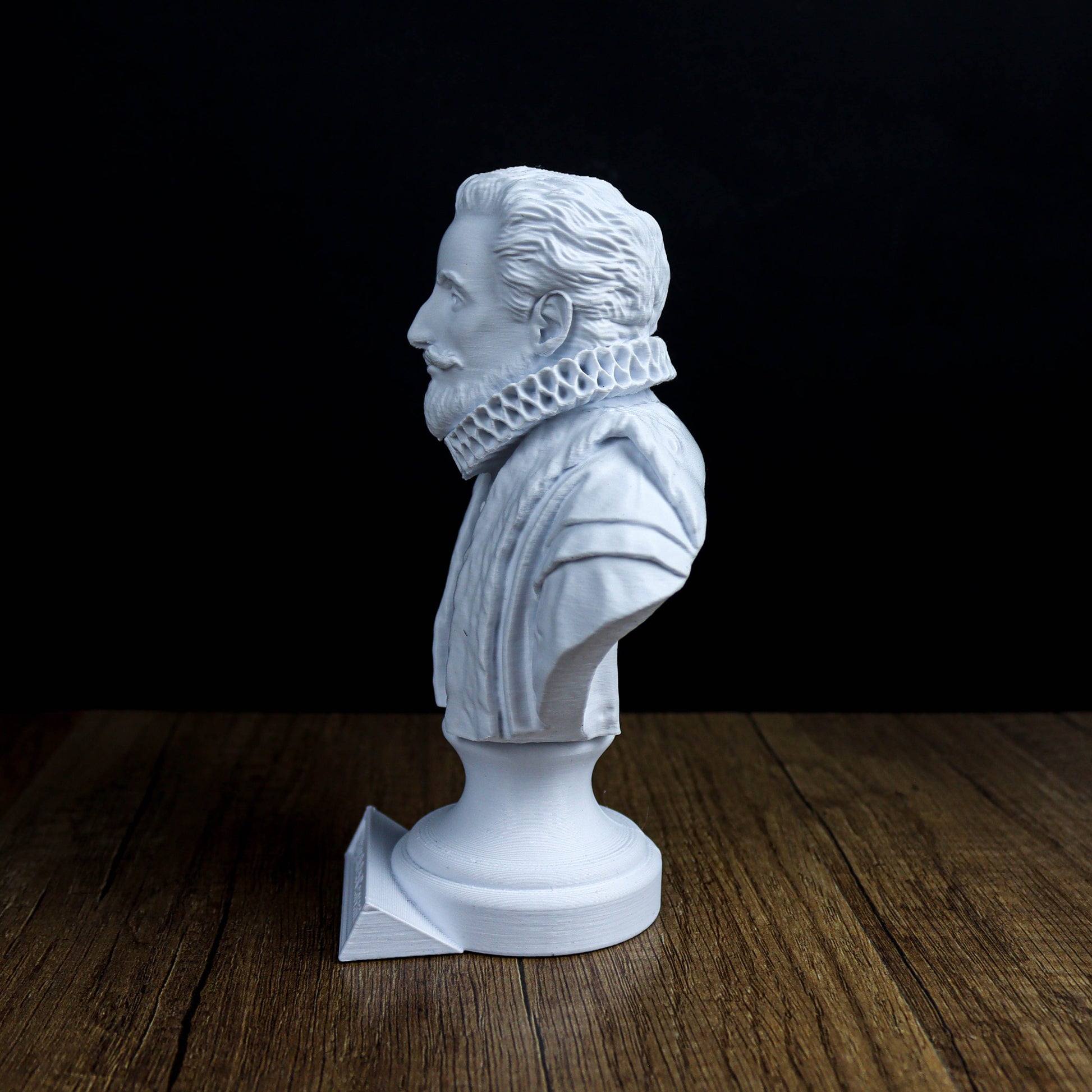 A white bust of a man with a beard and mustache, wearing a ruffled collar, is displayed on a wooden surface against a black background.