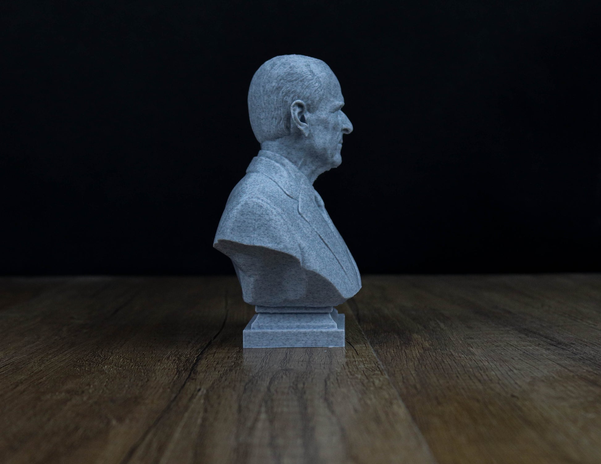 A gray bust of a man is displayed on a wooden surface against a dark background.