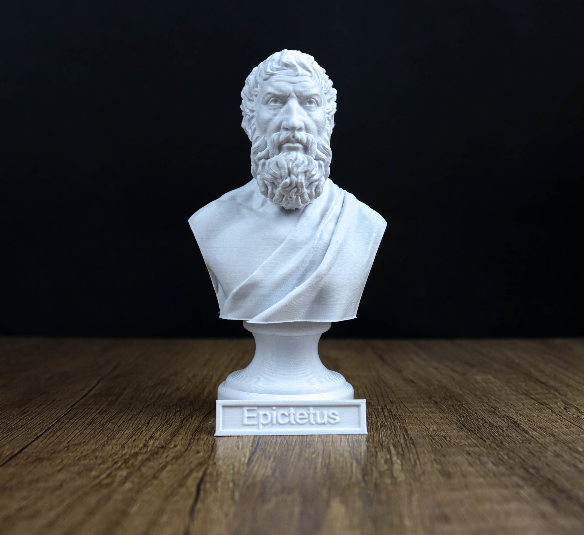 A white bust of a bearded man with a serious expression, likely depicting the ancient Greek philosopher Epicurus, is displayed on a wooden surface against a black background.