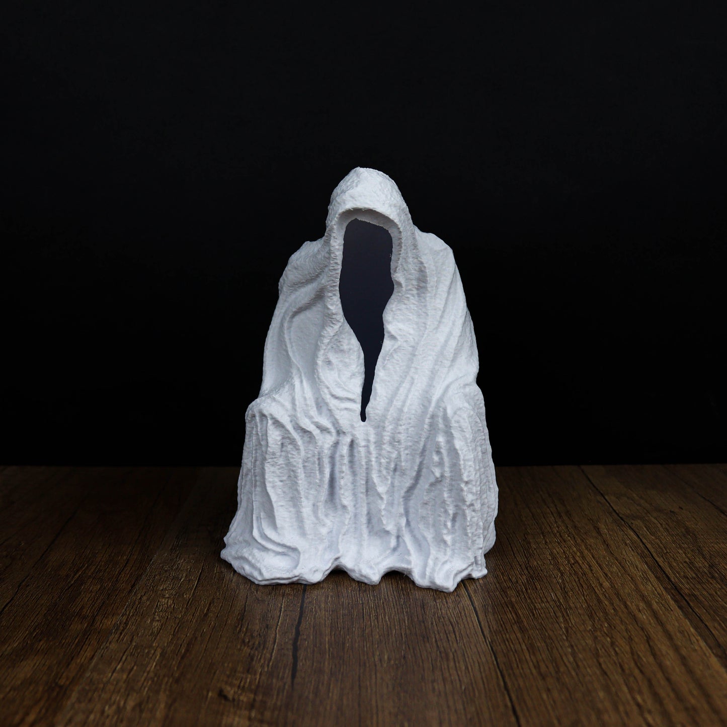 A white, hooded figure sits on a wooden surface against a dark background.