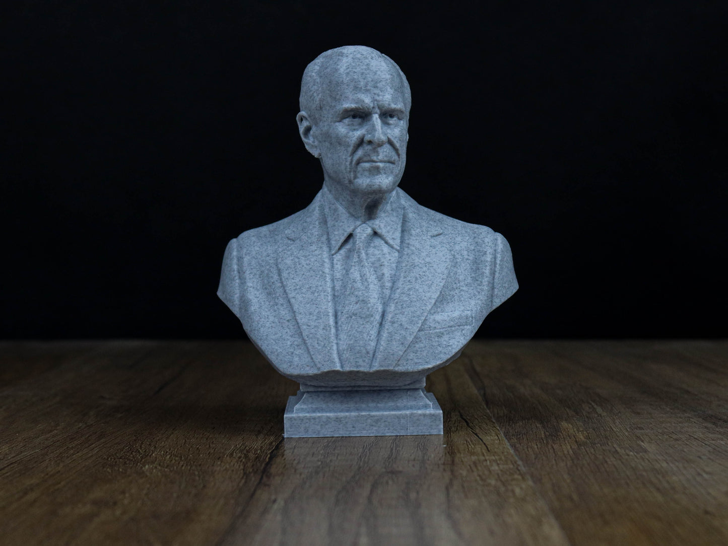 A gray stone bust of a man wearing a suit and tie, sitting on a wooden surface against a dark background.