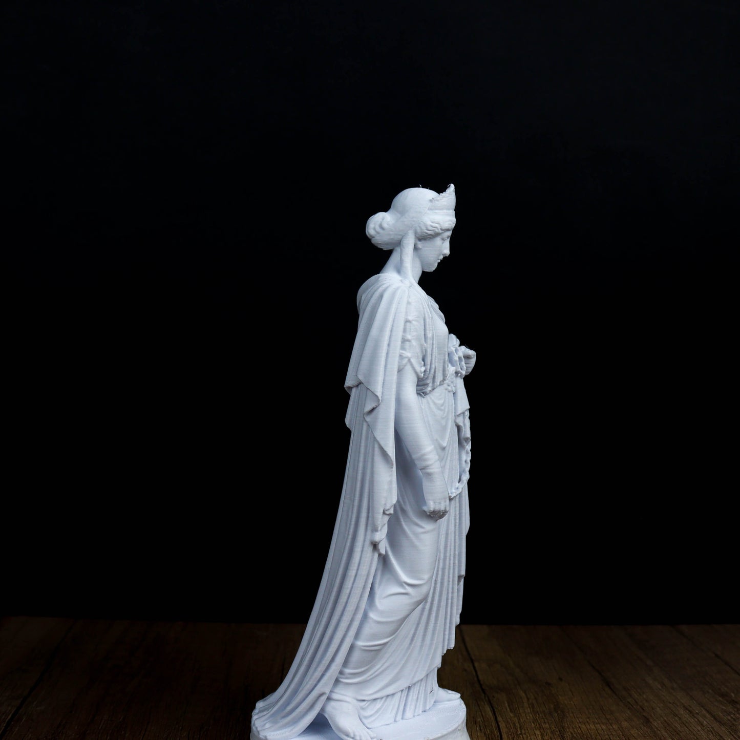 A white marble statue of a woman with a flowing robe and headdress stands on a wooden surface against a dark background.