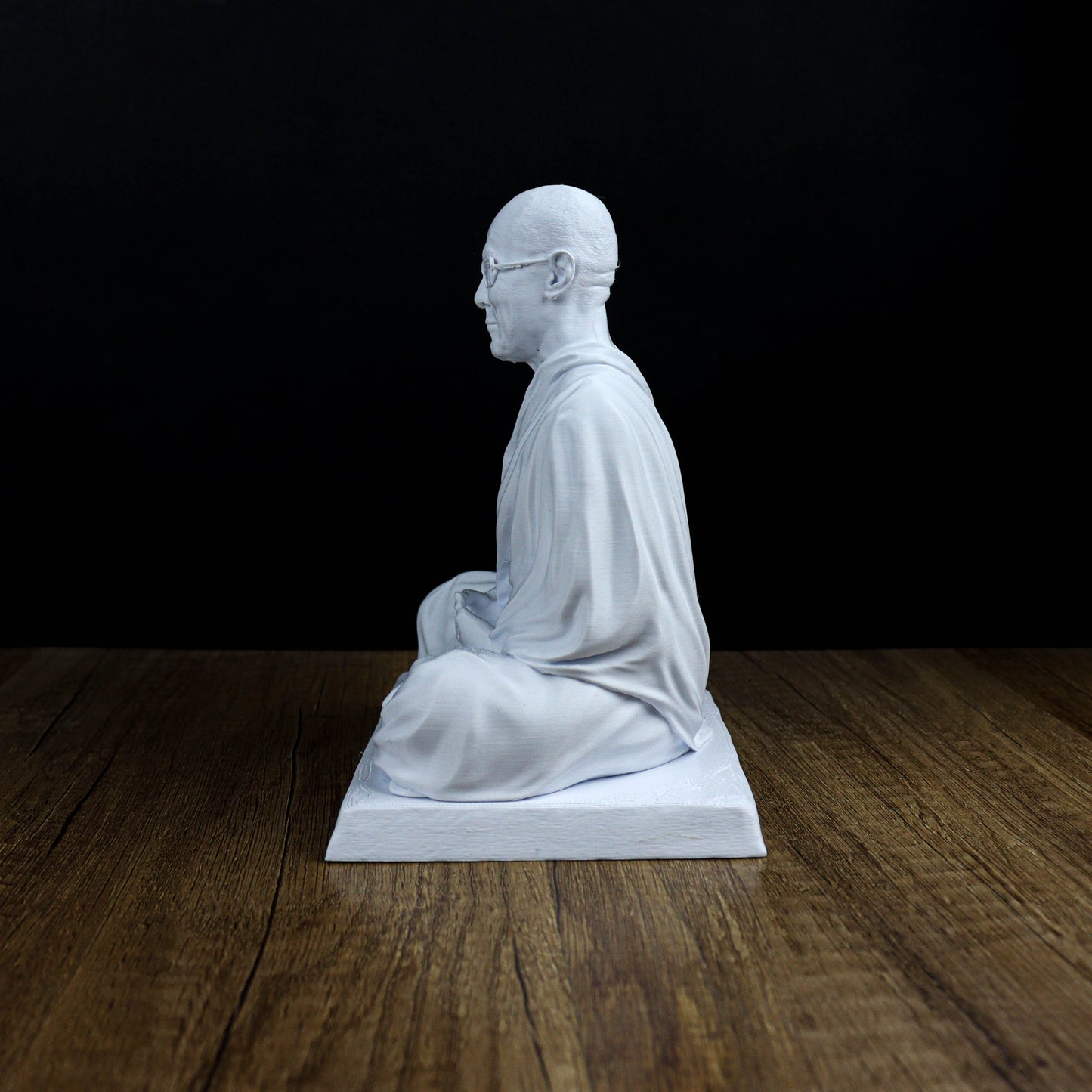 A white statue of a seated Buddha figure is displayed on a wooden surface against a black background.