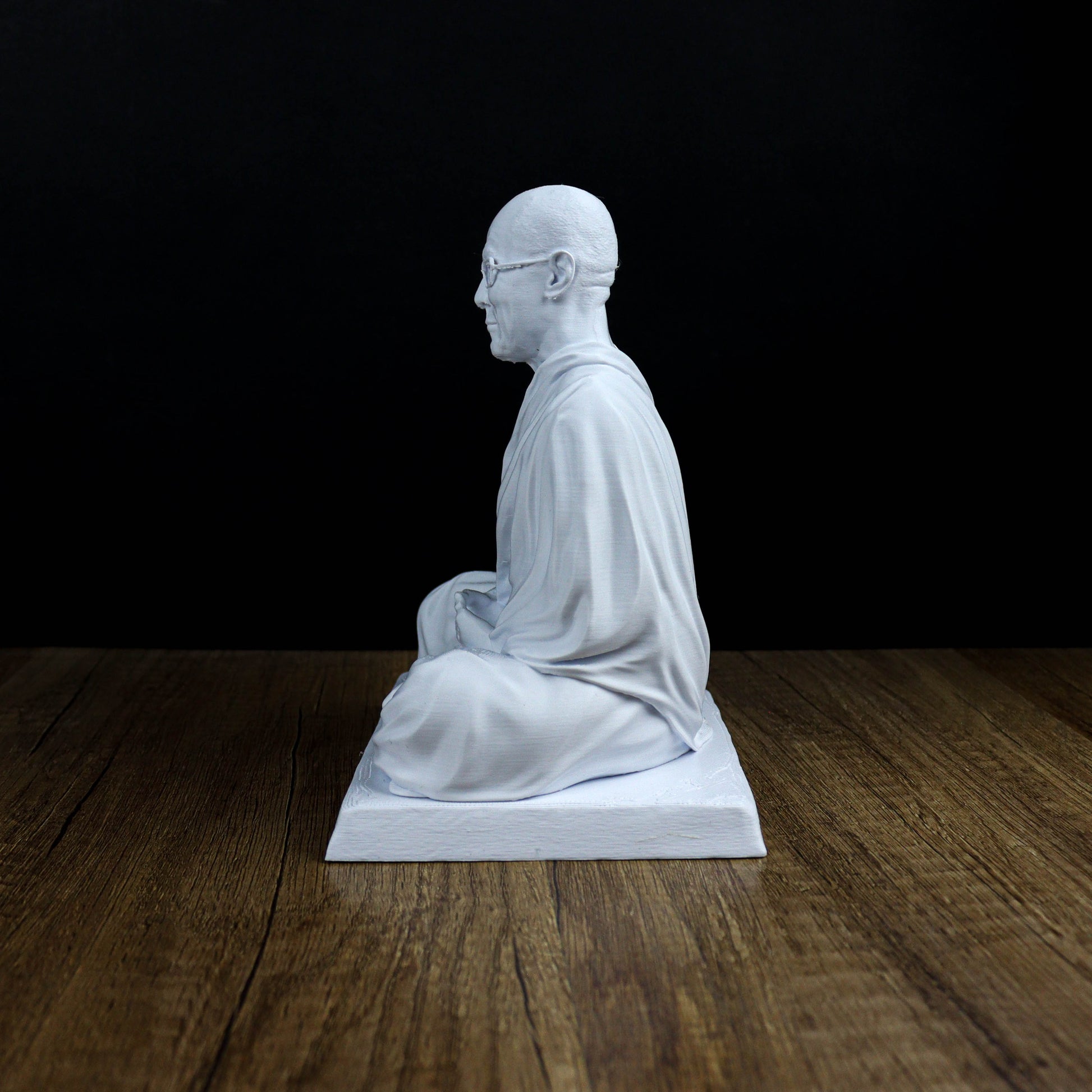 A white statue of a seated Buddha figure is displayed on a wooden surface against a black background.