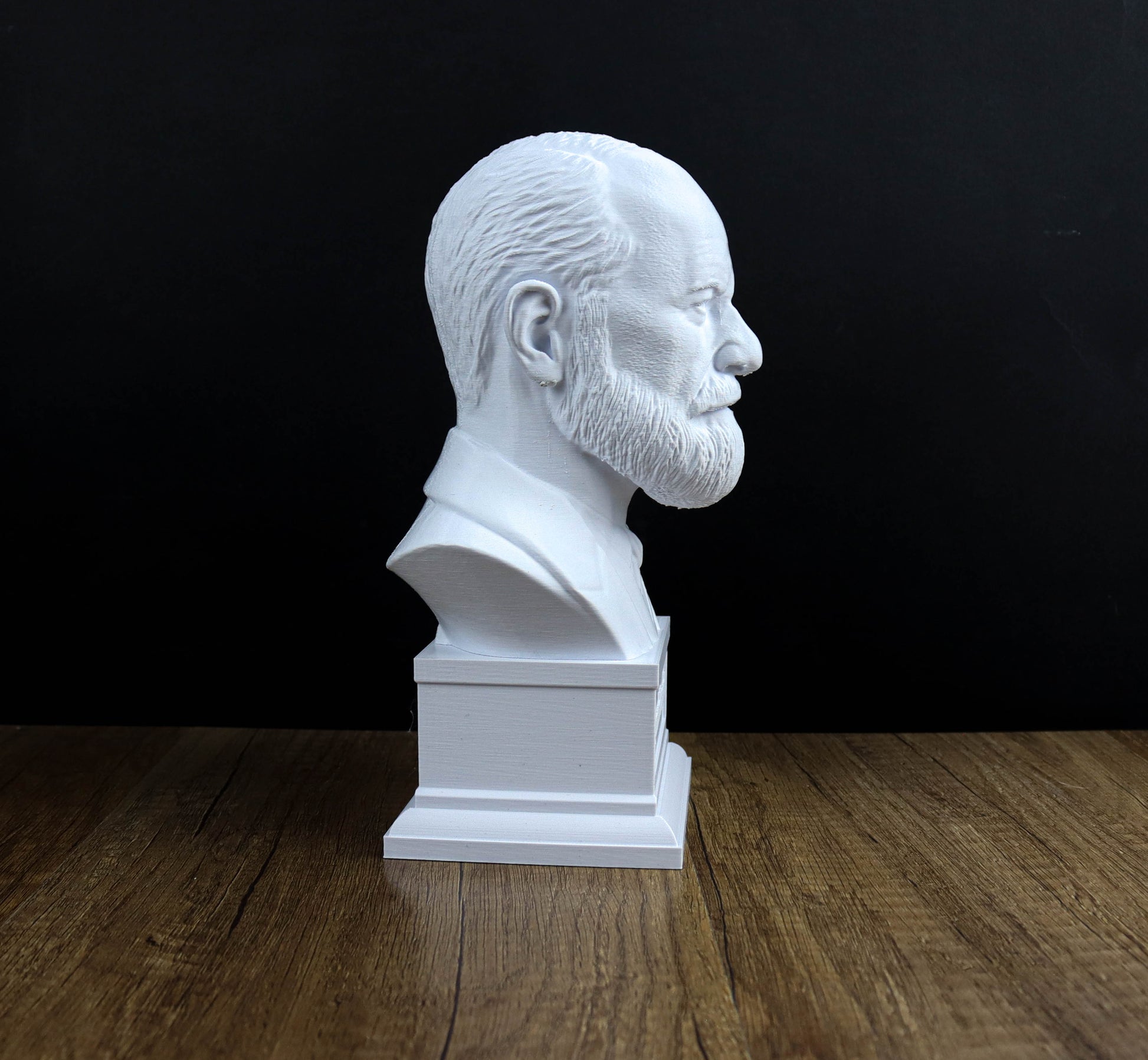 A white bust of a man with a beard is displayed on a wooden surface against a black background.