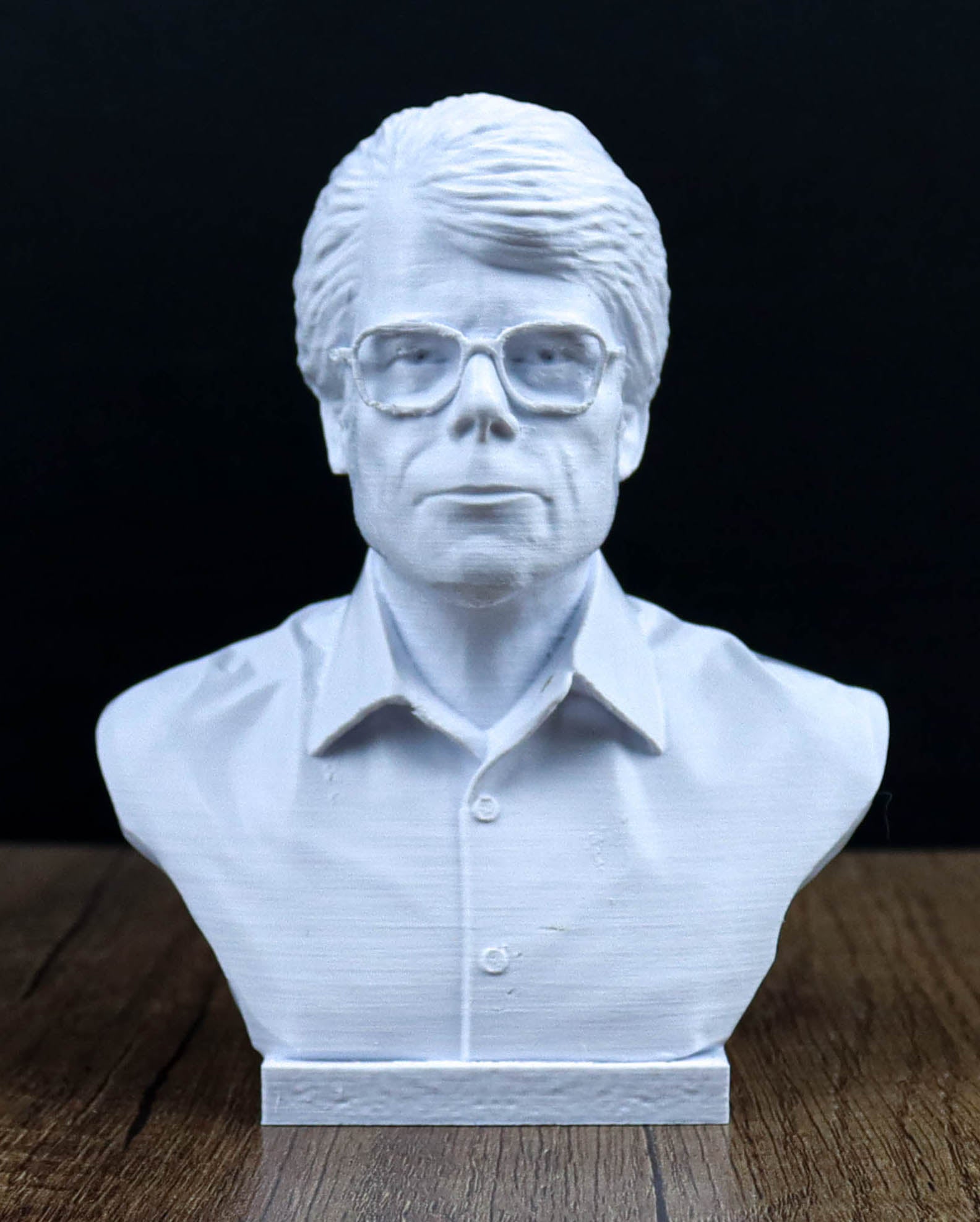 A white bust sculpture of a man wearing glasses, placed on a wooden surface against a dark background.