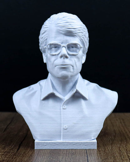 A white bust sculpture of a man wearing glasses, placed on a wooden surface against a dark background.