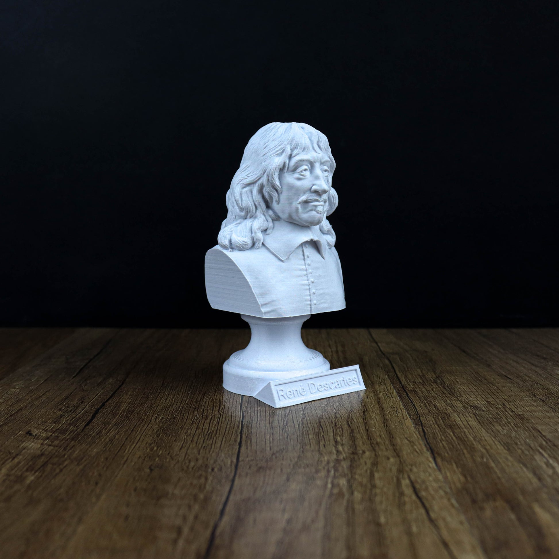 A white bust of a man with long hair and a mustache is displayed on a wooden surface against a black background.