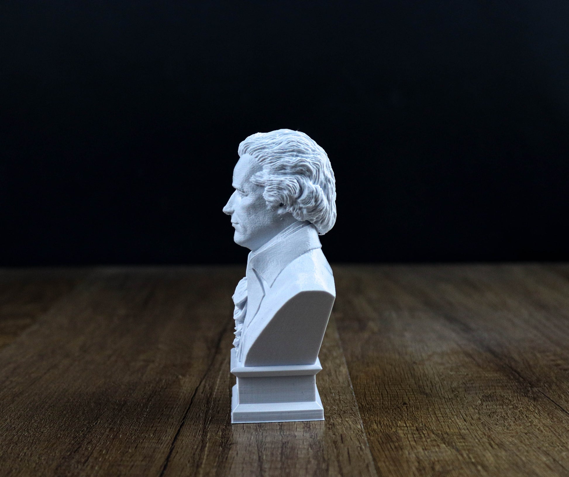 A white bust of a classical musician is displayed on a wooden surface against a dark background.
