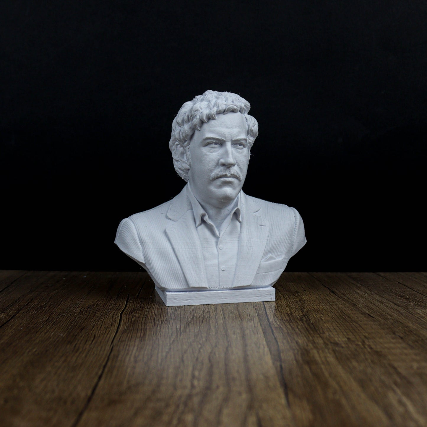 A white bust of a man with curly hair and a mustache, wearing a suit, is displayed on a wooden surface against a dark background.
