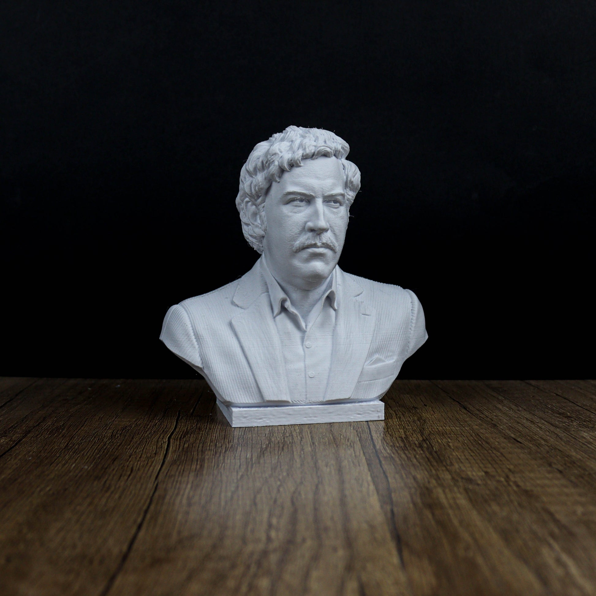 A white bust of a man with curly hair and a mustache, wearing a suit, is displayed on a wooden surface against a dark background.
