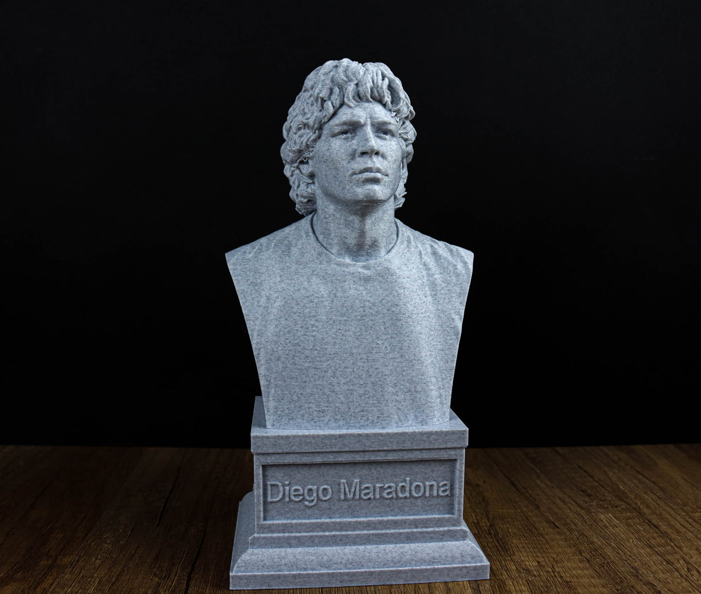 A bust of Diego Maradona, a famous soccer player, is displayed on a wooden surface against a black background.