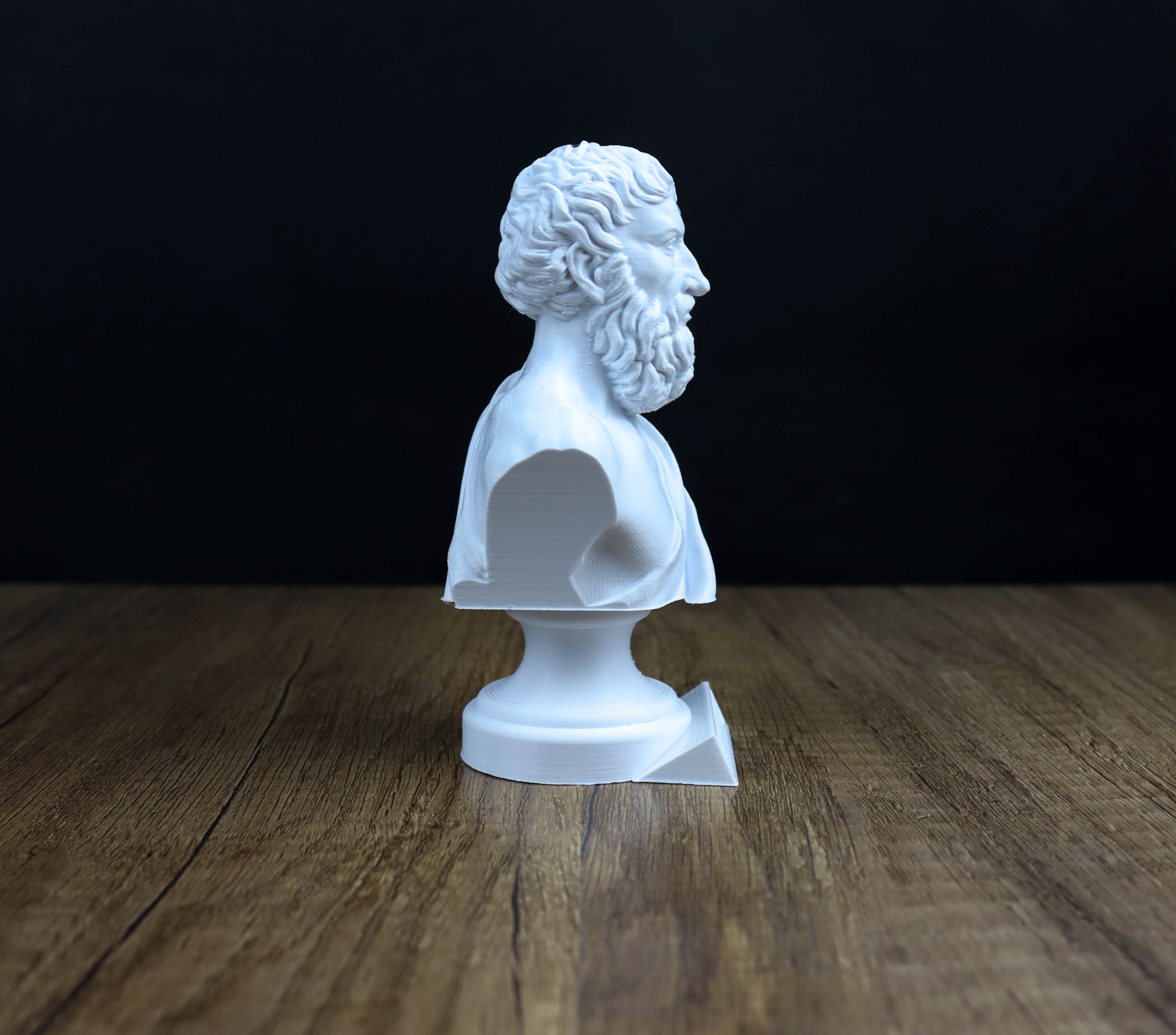 A white bust of a bearded man is displayed on a wooden surface against a dark background.