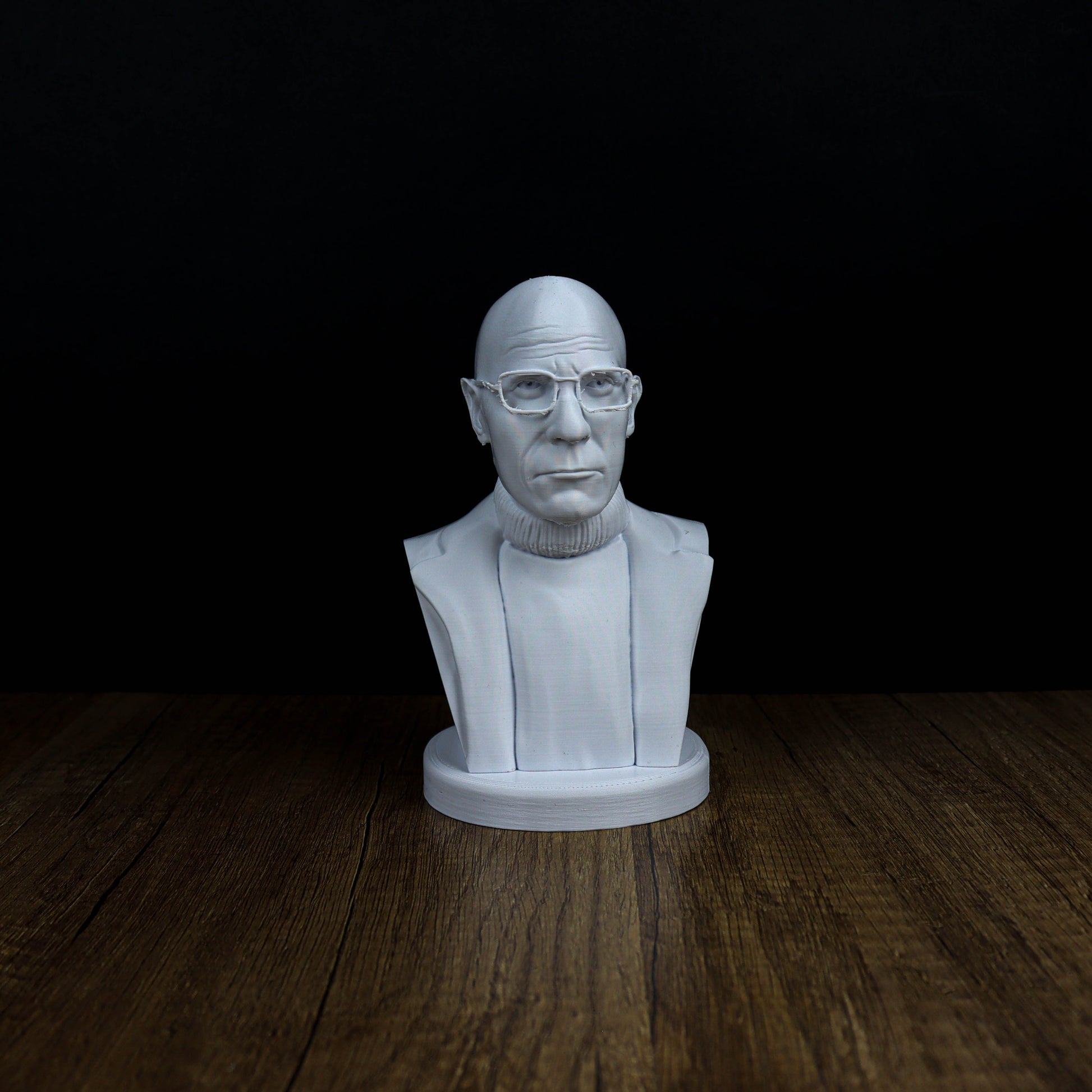 A white bust of a man wearing glasses is displayed on a wooden surface against a black background.