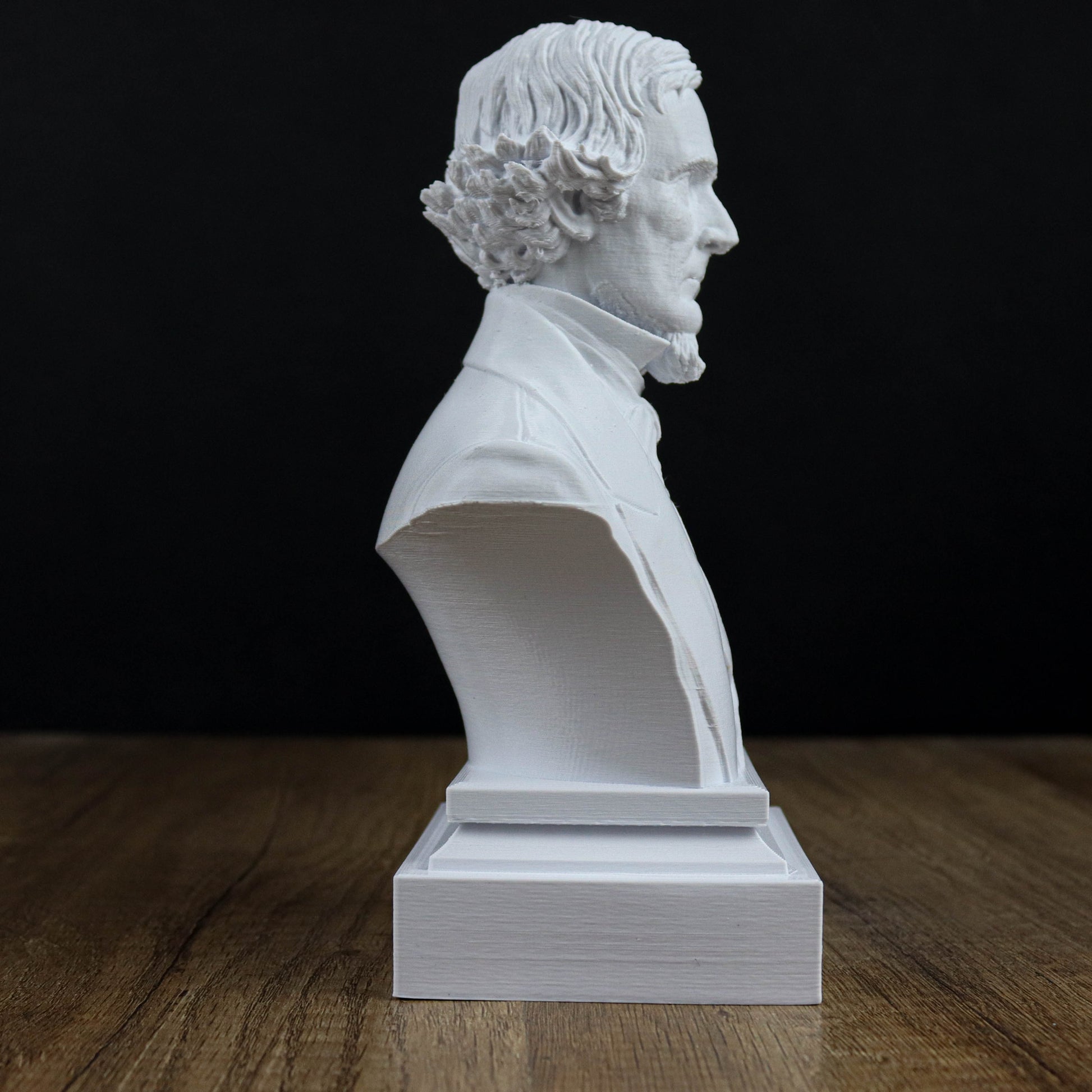 A white bust of a man with curly hair and a beard is displayed on a wooden surface against a black background.