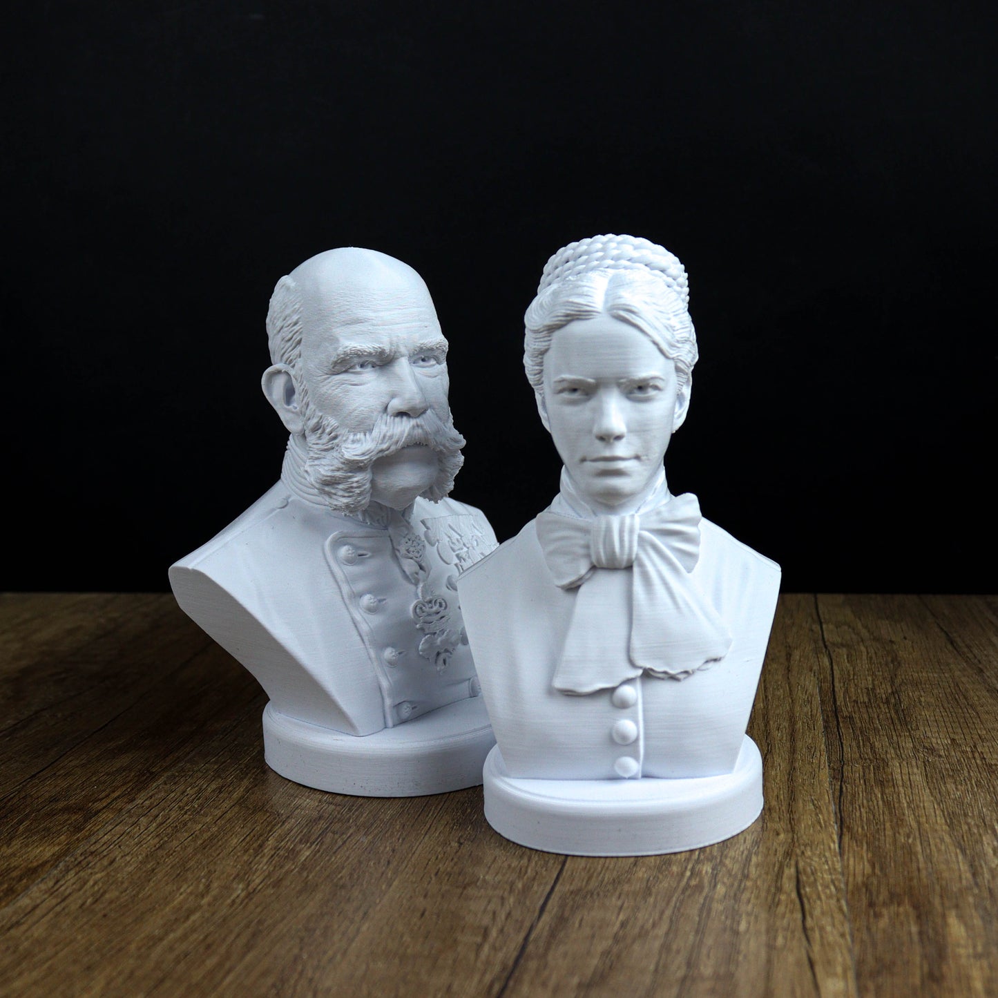 Two white busts of a man and a woman, both with distinct hairstyles and clothing, are displayed on a wooden surface against a black background.