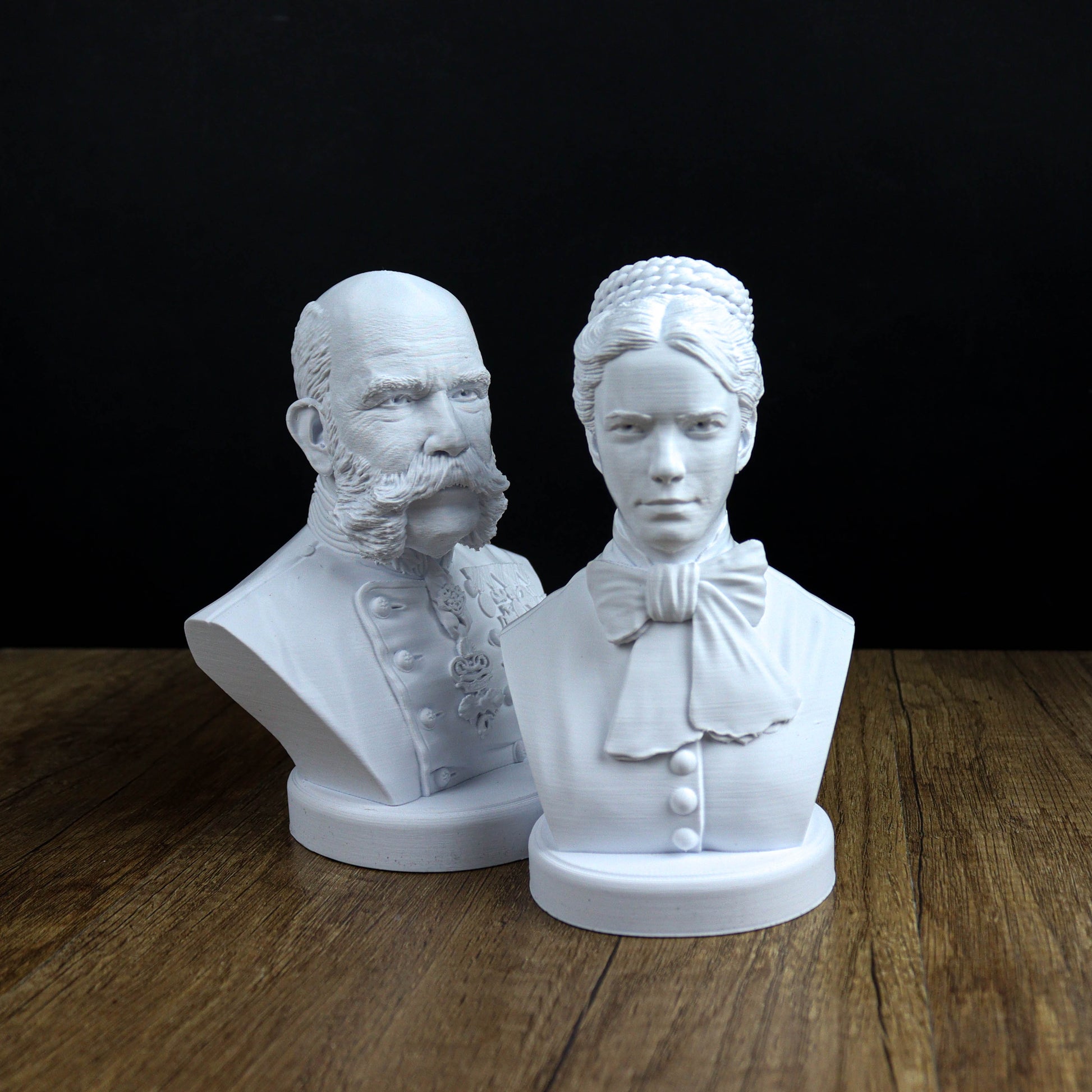 Two white busts of a man and a woman, both with distinct hairstyles and clothing, are displayed on a wooden surface against a black background.