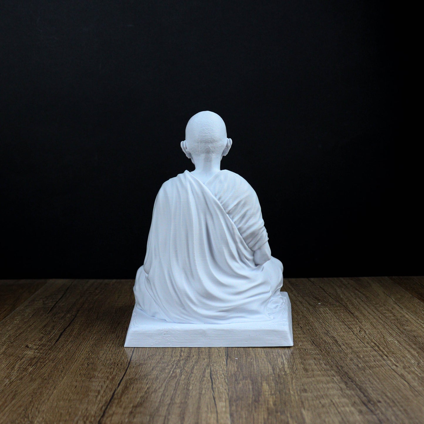 A white statue of a seated Buddha figure is displayed on a wooden surface against a black background.