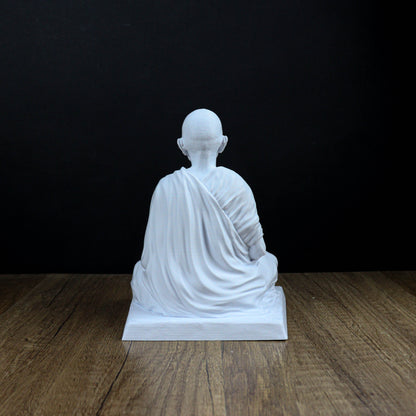 A white statue of a seated Buddha figure is displayed on a wooden surface against a black background.